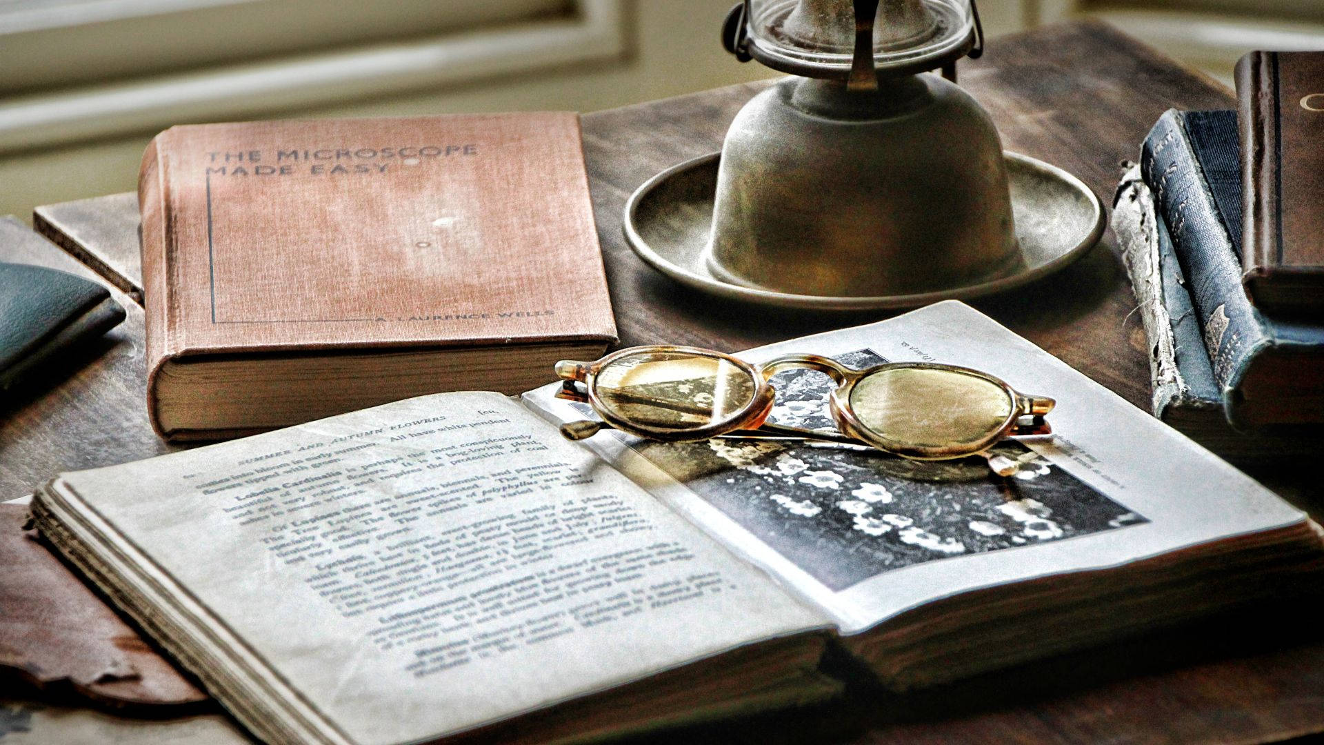 glasses 桌布 壁紙 — 一本打開的復古書本連同老花眼鏡，放在木桌上，旁邊有一盞黃銅燈和舊書。
