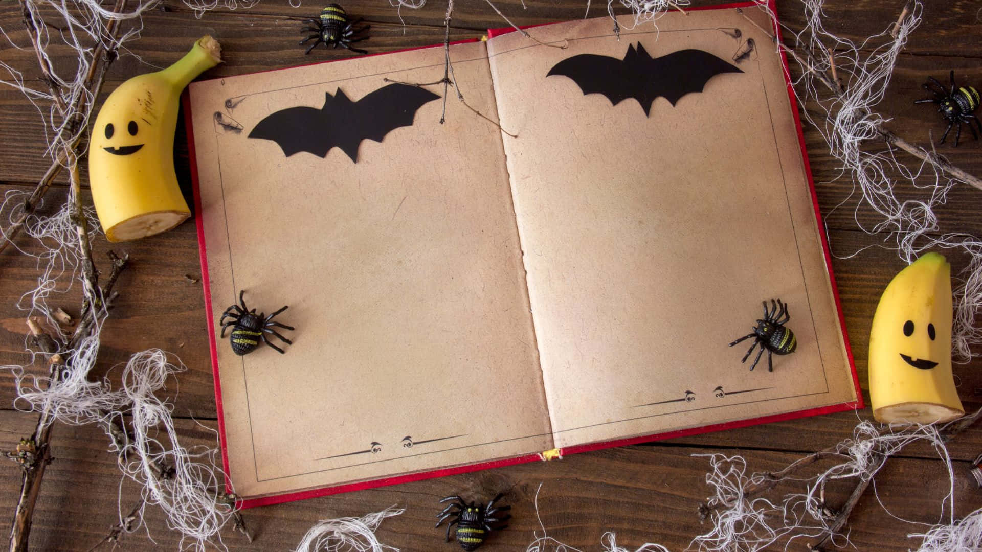 Halloween Books 桌布 壁紙 — 一本打開的書，上面有蝙蝠剪紙和微笑的香蕉半邊，周圍是假蜘蛛和蜘蛛網，放在木桌上。