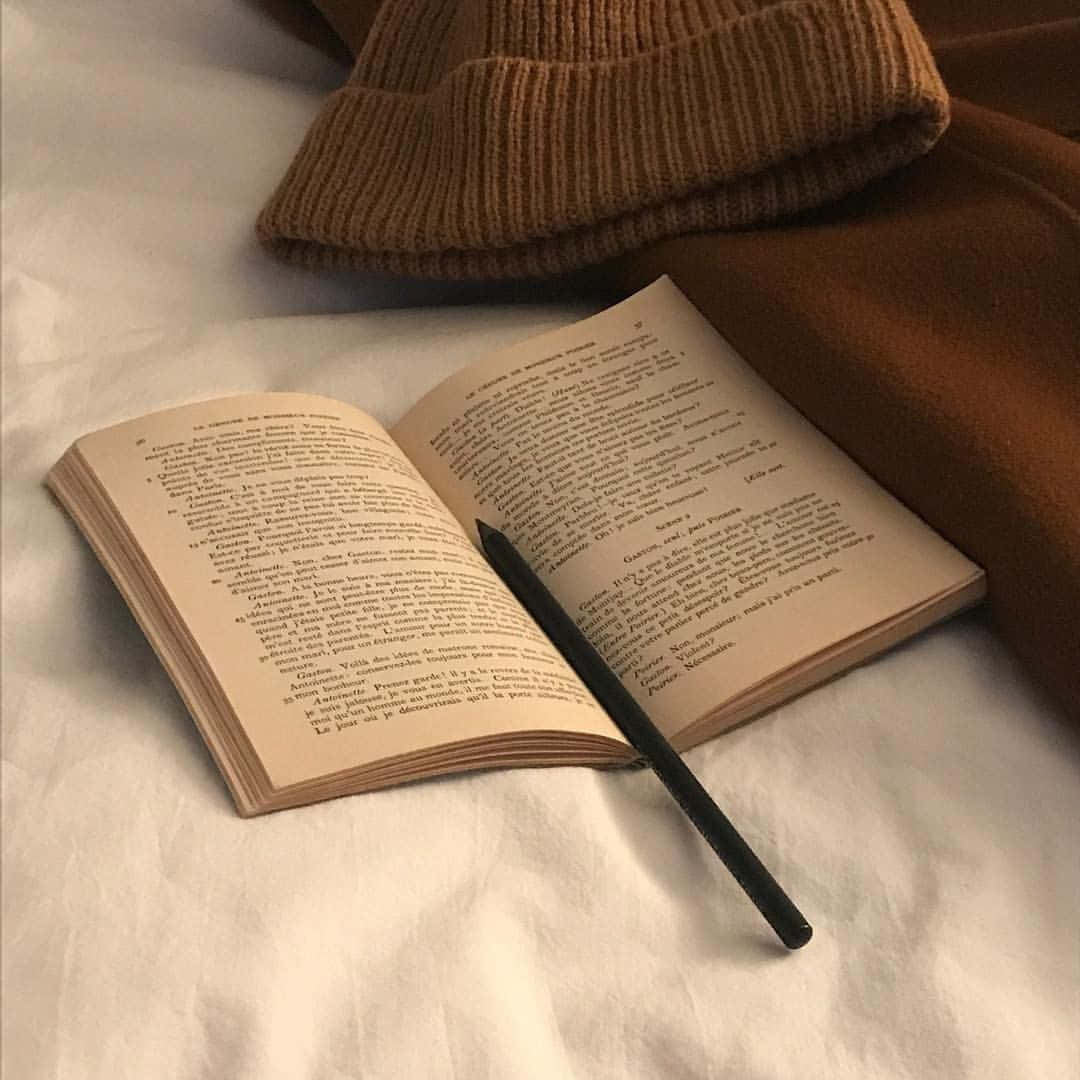 棕色美學 桌布 壁紙 — 一本打開的書，旁邊有一支黑色筆，還有一頂舒適的棕色針織帽，放在柔軟的白色床罩上。