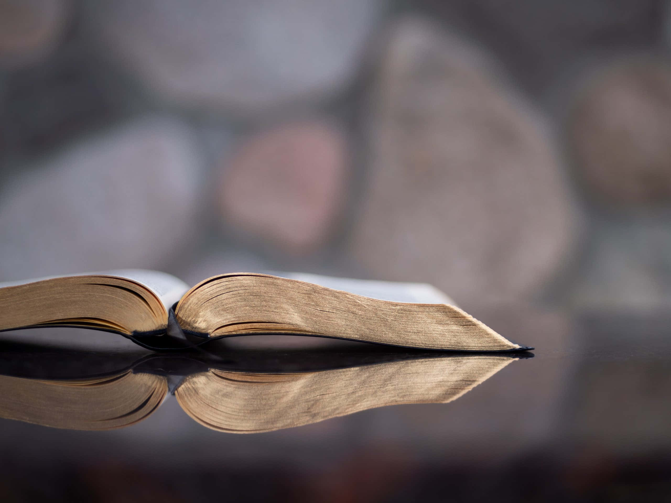 Scripture 桌布 壁紙 — 一本打開的書，泛黃的書頁放在反光表面上，背景是模糊的石牆。