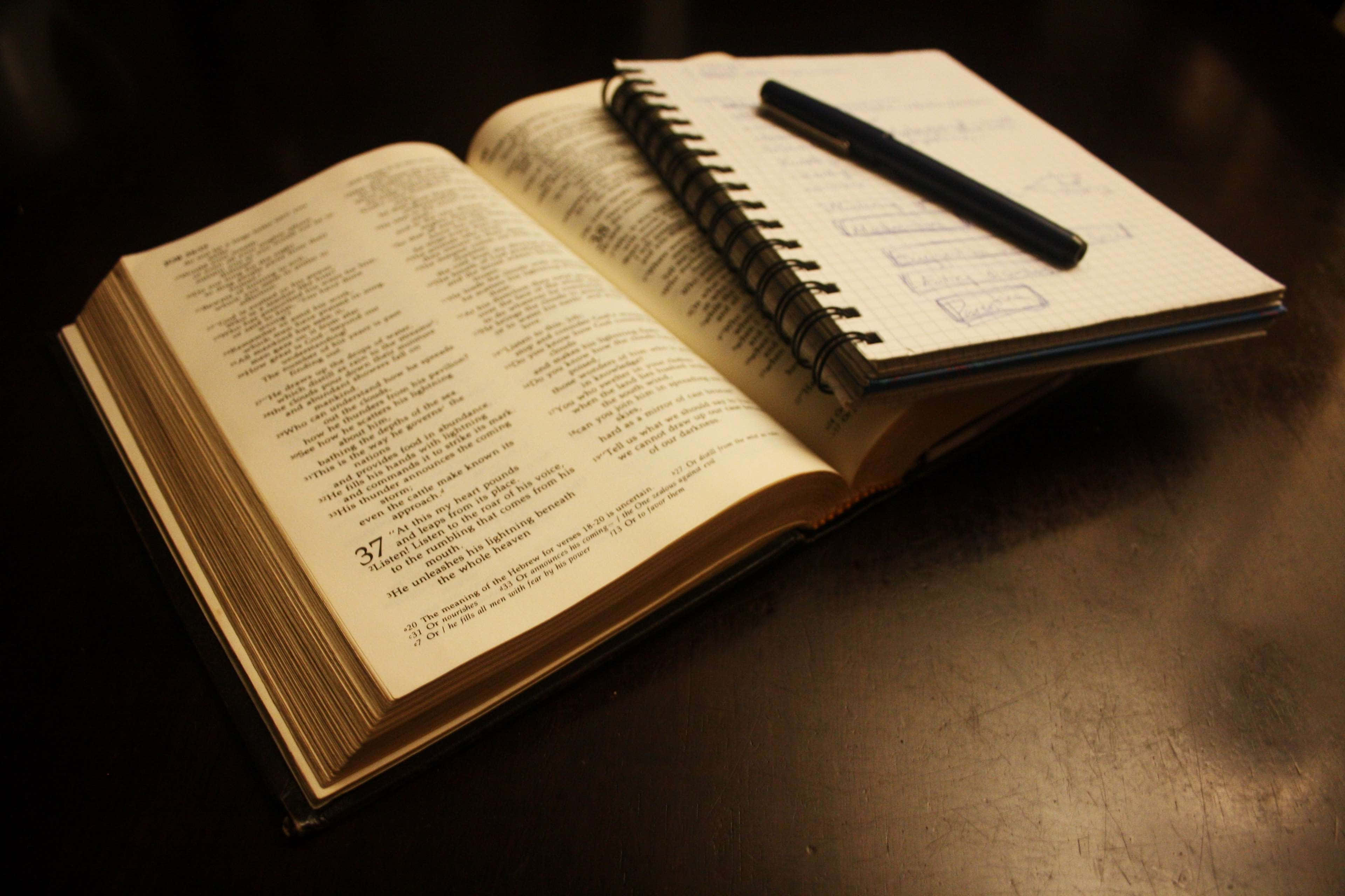 Bible Study 桌布 壁紙 — 一本打開的書，連同螺旋筆記本和筆，放在深色木桌上，溫暖的環境光下。