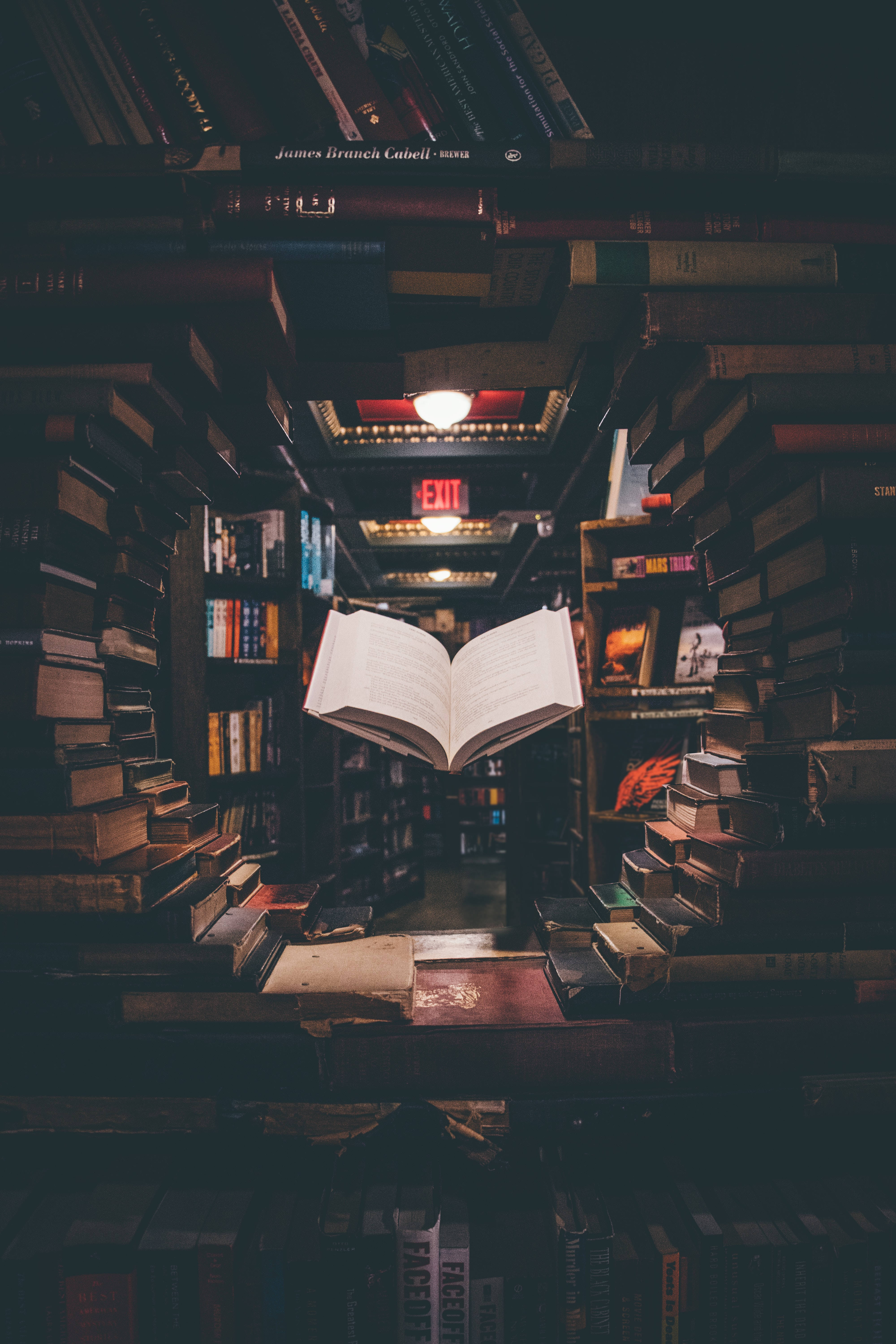 書本 桌布 壁紙 — 一本打開的書在燈光昏暗的書店通道中神秘地漂浮，周圍是高聳的書架。