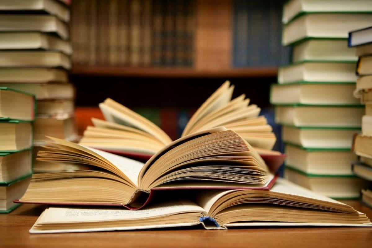 書本 桌布 壁紙 — 一本打開的書放在木桌上，周圍是堆疊如山的書籍，營造出舒適的圖書館氛圍。
