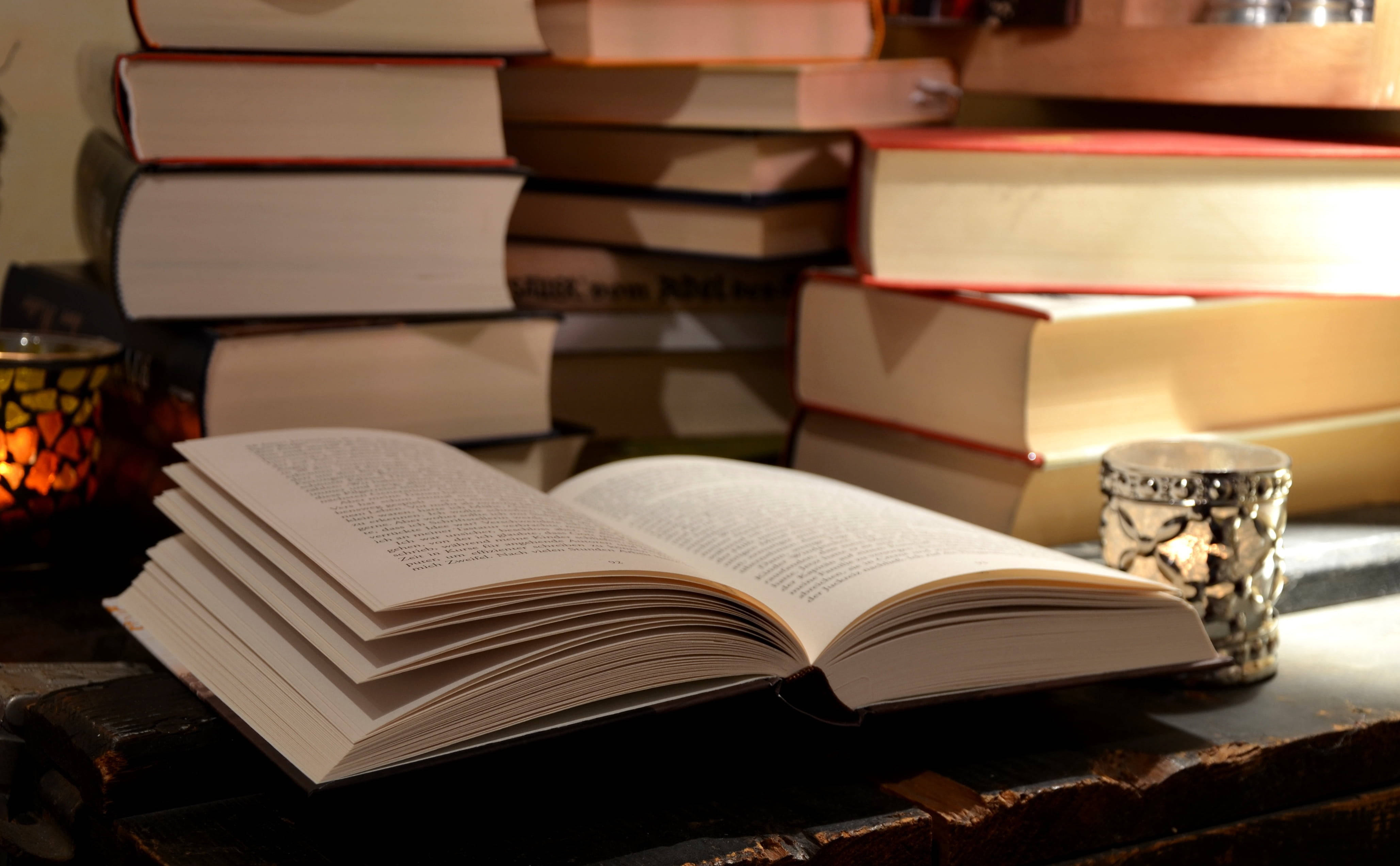Reading 桌布 壁紙 — 一本打開的書放在木桌上，周圍是成堆的書籍和發光的蠟燭，在溫暖的環境光中。