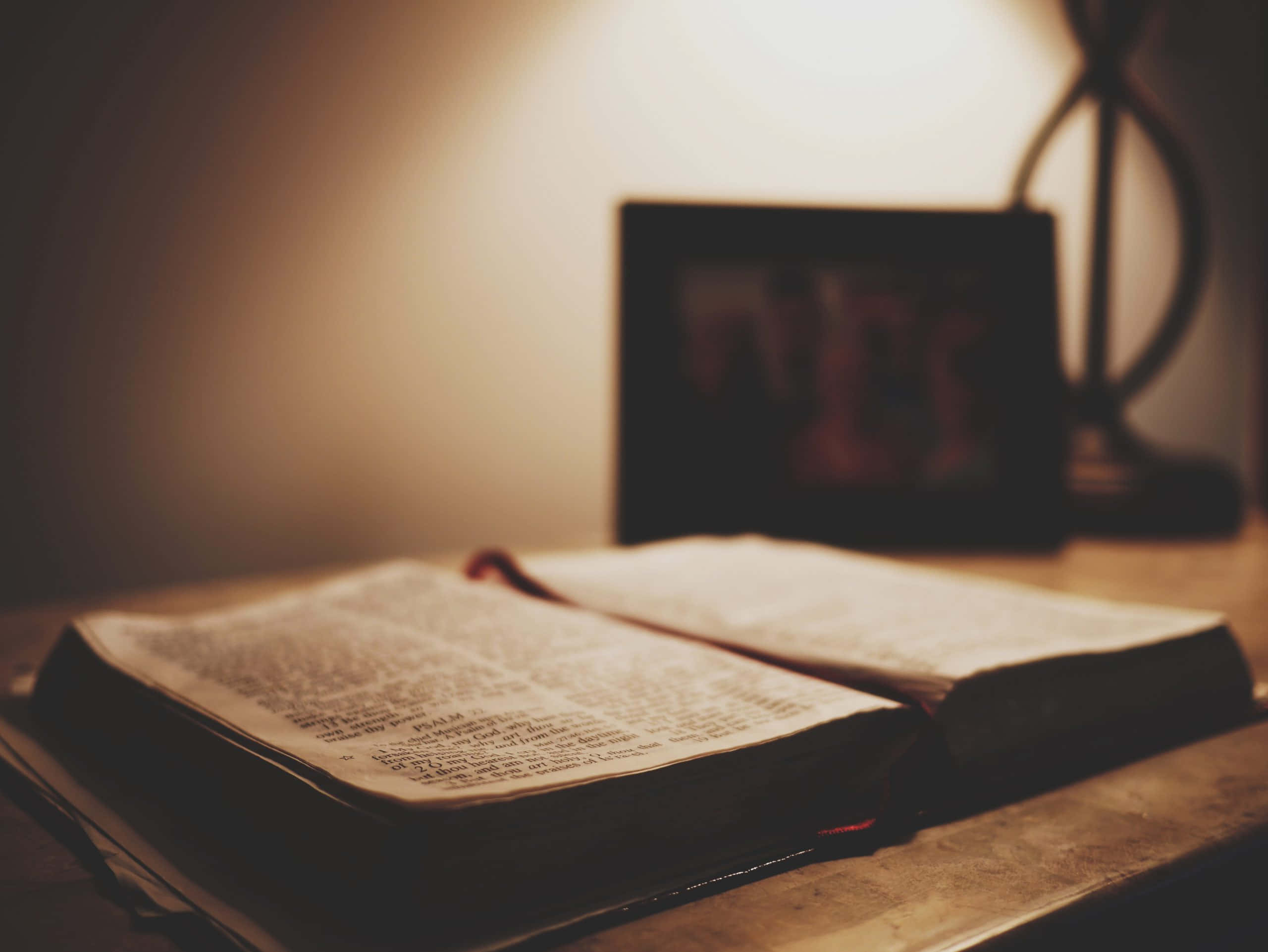 Scripture 桌布 壁紙 — 一本打開的書放在木桌上，沐浴在溫暖的燈光中，背景中有一個裱框的照片柔和地模糊。