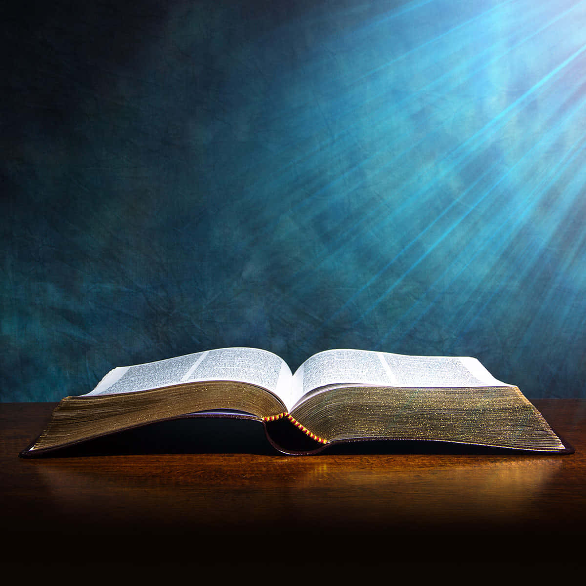聖經 桌布 壁紙 — 一本打開的書放在木質表面上，神聖的光芒從上方照射下來，映襯著深藍色紋理背景。
