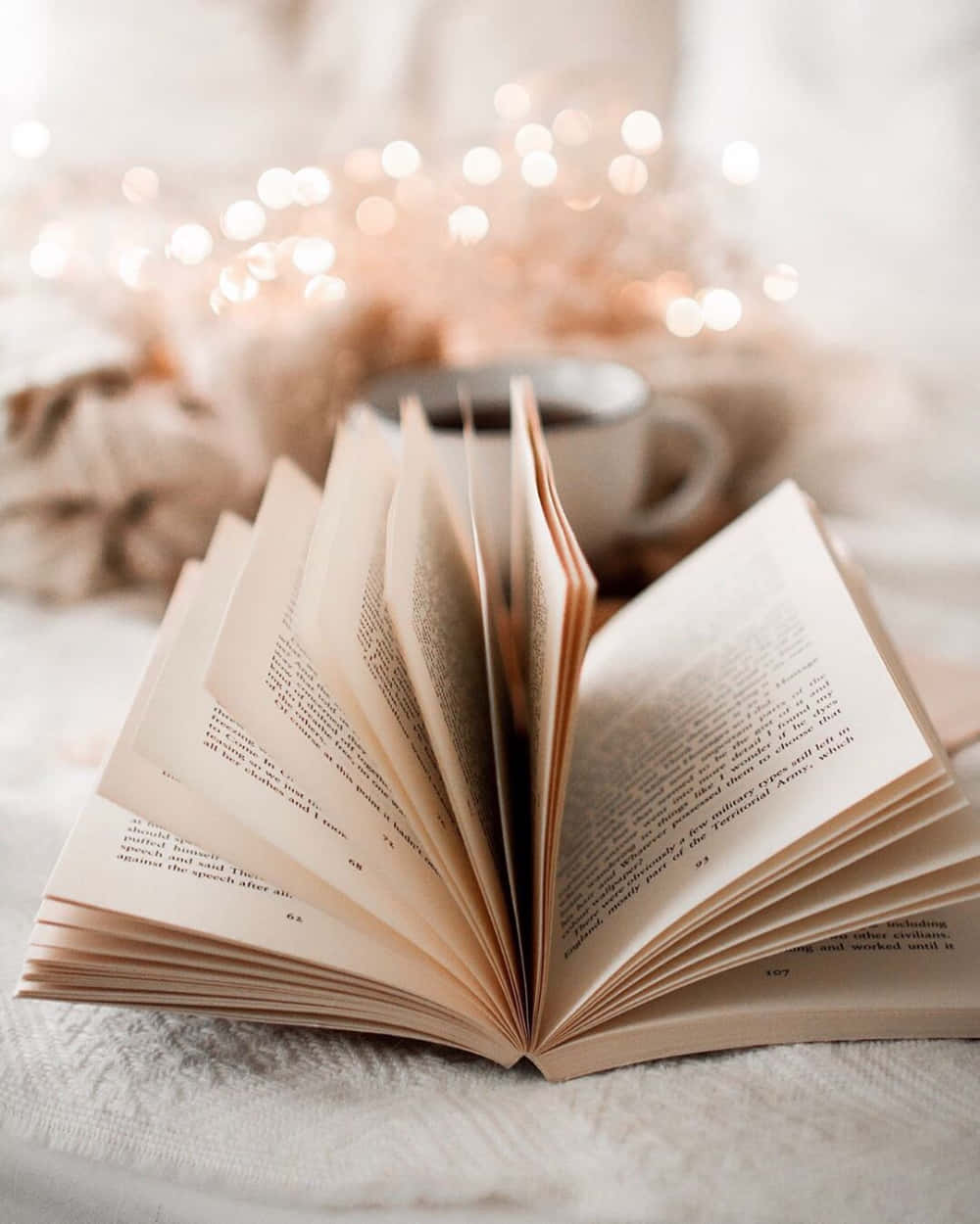 Open Book 桌布 壁紙 — 一本打開的書放在柔軟有質感的表面上，旁邊有舒適的杯子和溫暖的仙女燈在背景中柔和地發光。