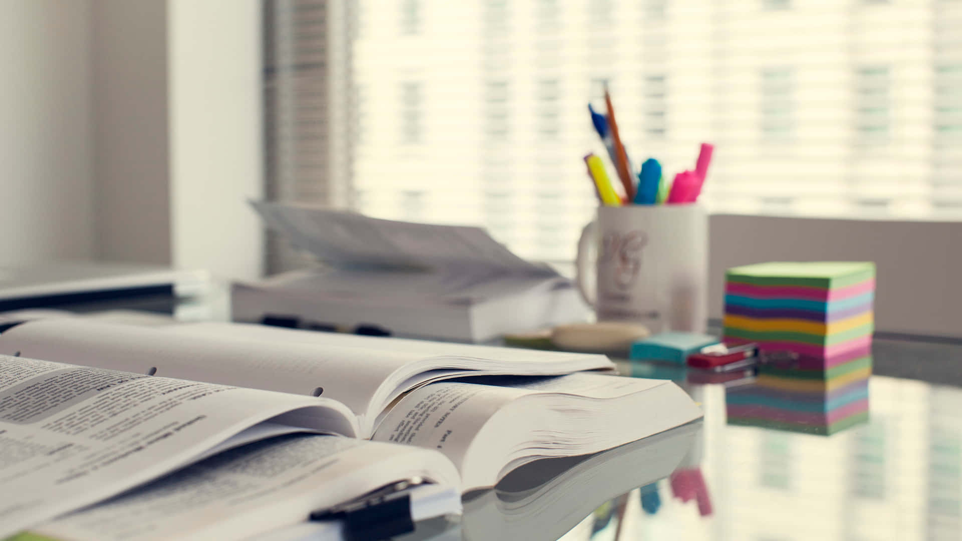 Desk 桌布 壁紙 — 一本打開的書放在玻璃桌上，旁邊有彩色便利貼和一個裝滿筆的杯子，靠近帶百葉窗的窗戶。