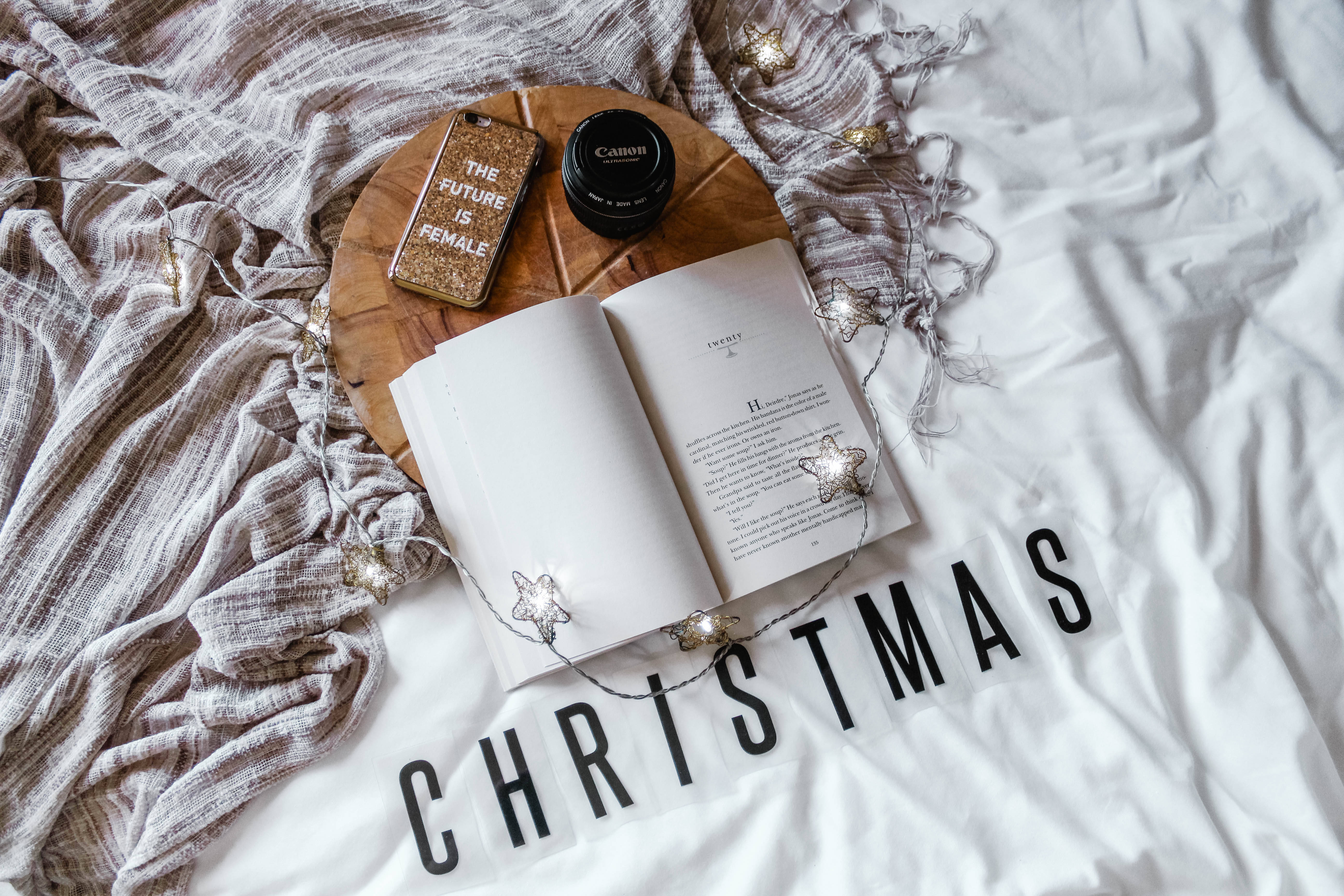 聖誕節手機 桌布 壁紙 — 一本打開的書放在舒適的床上，'CHRISTMAS' 以粗體字母拼出，周圍環繞著節日燈光和閃亮的手機殼。