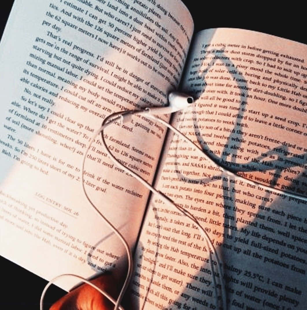 Open Book 桌布 壁紙 — 一本打開的書的特寫，白色耳塞放在書頁上，沐浴在溫暖的陽光中。
