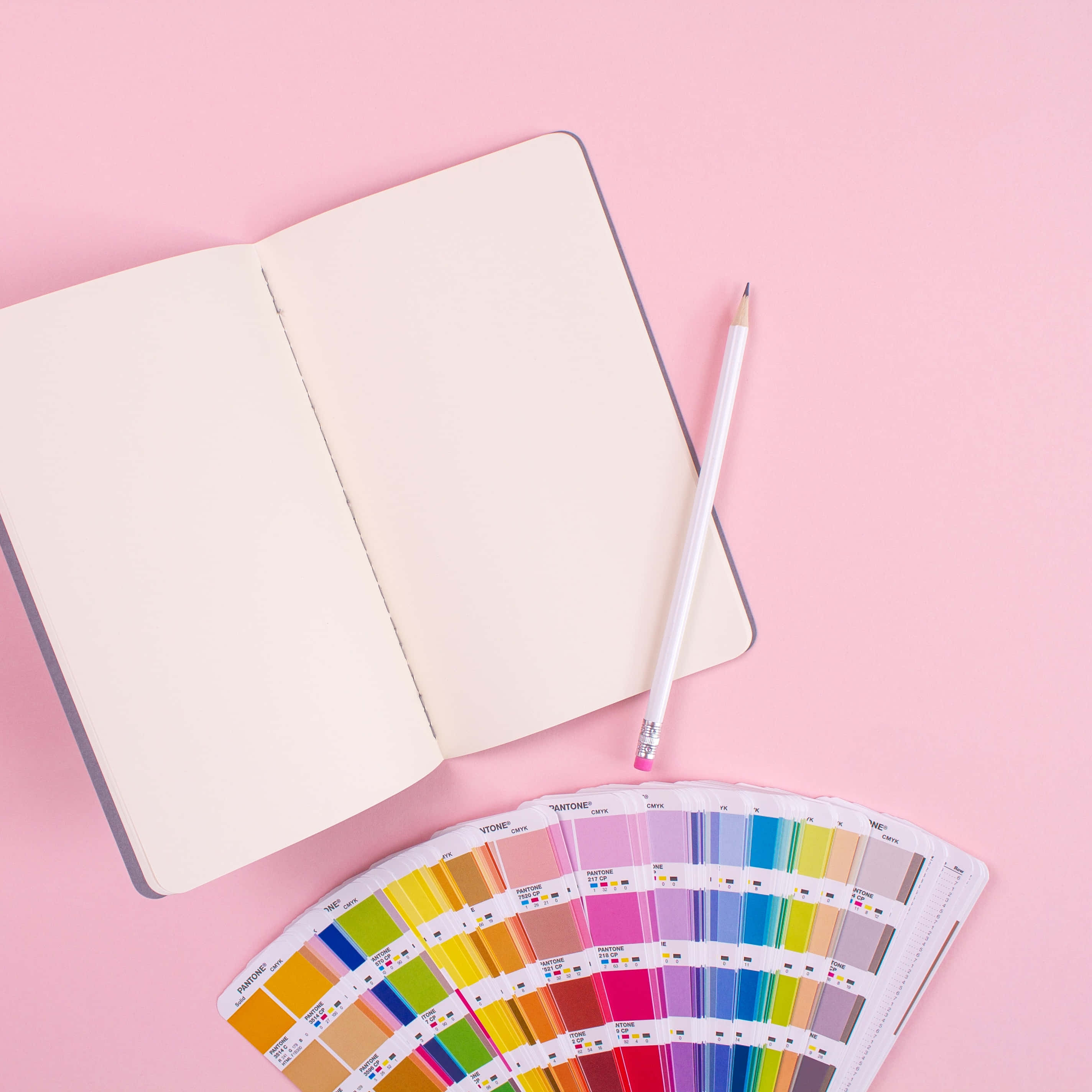 Notebook 桌布 壁紙 — 一本打開的空白筆記本和鉛筆放在一個展開的 Pantone 色票扇旁邊，背景是柔和的粉紅色。