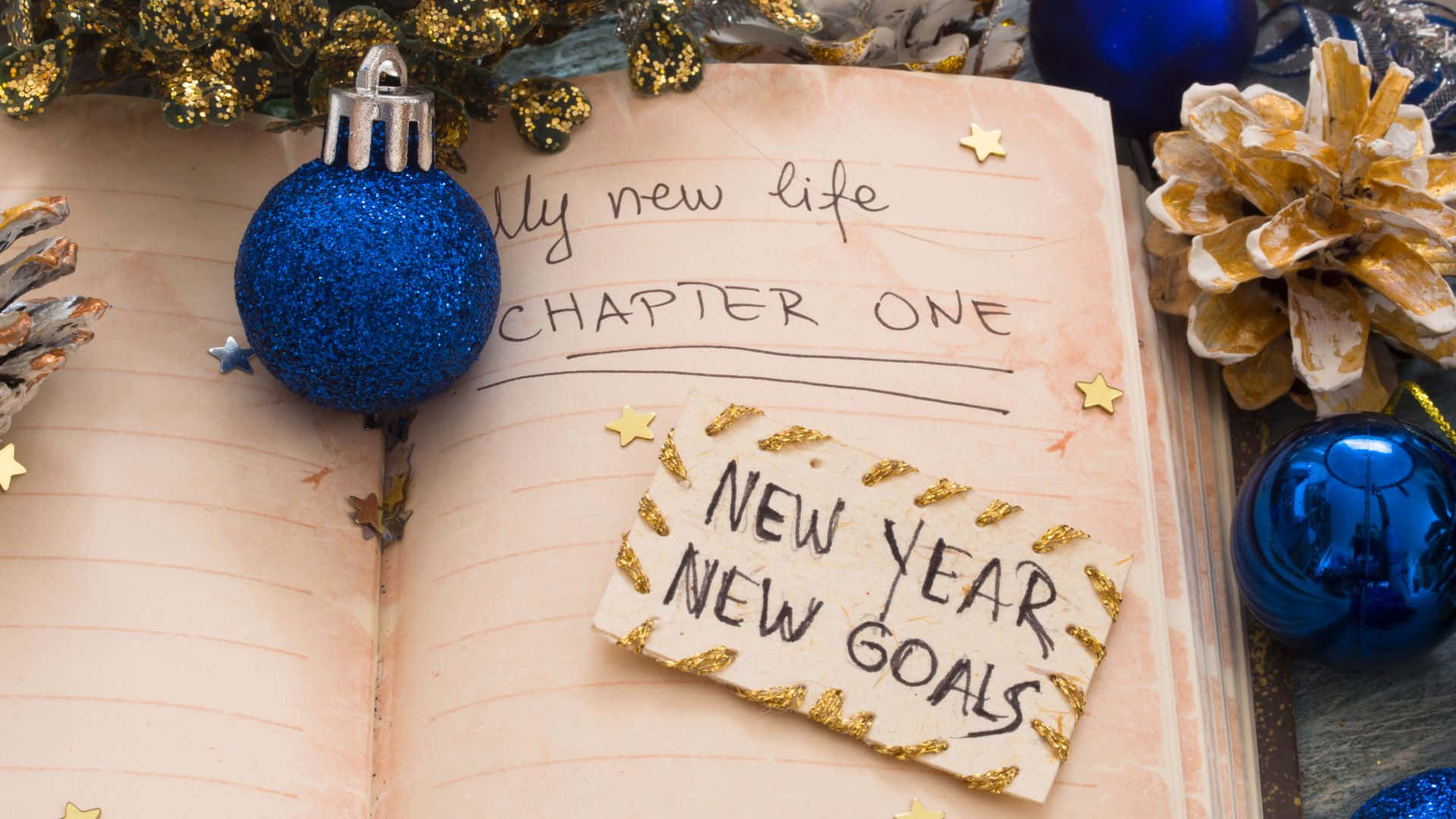 桌布 壁紙 — 一本打開的筆記本，手寫著「CHAPTER ONE」和「NEW YEAR NEW GOALS」，周圍環繞著藍色亮粉裝飾、松果和金色星星。