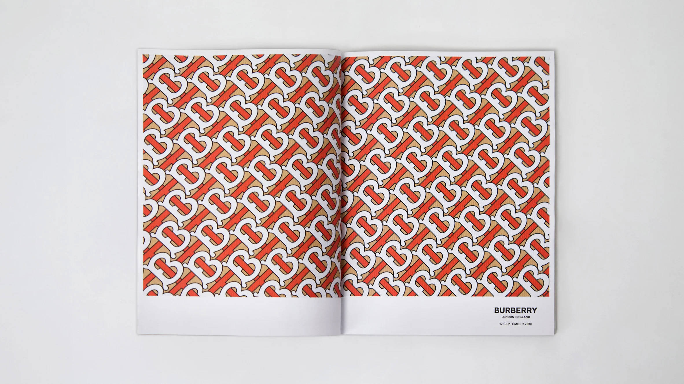 Burberry 桌布 壁紙 — 一本打開的雜誌在純白色背景上展示米色頁面上重複的紅白交錯「B」標誌大膽圖案。