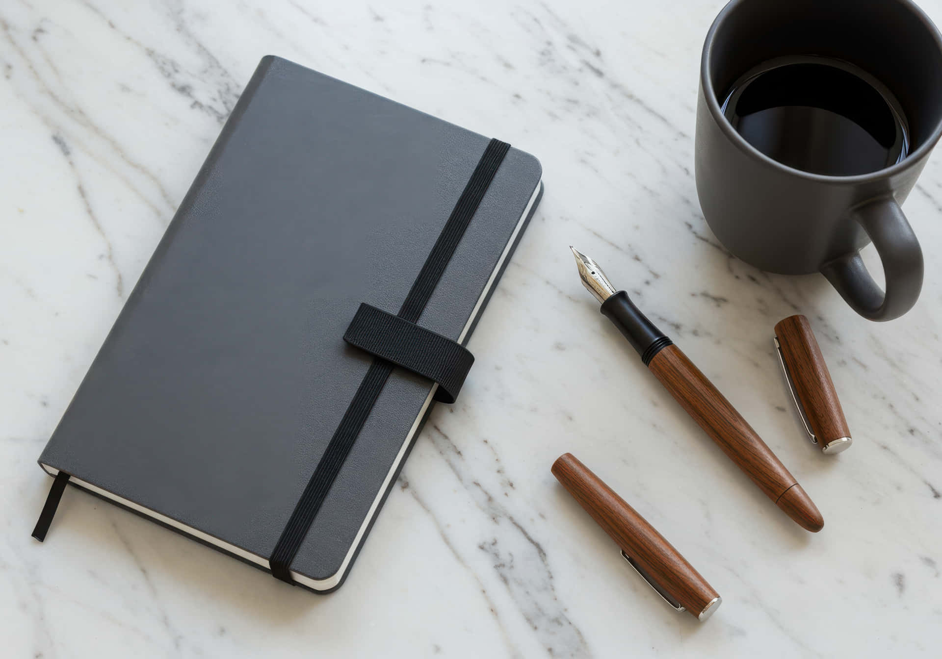 Gray notebook with a pen and coffee mug on a marble surface 壁紙 — 一本時尚的灰色筆記本，帶有黑色彈性帶，旁邊放著一支木質鋼筆和一個深色咖啡杯，置於白色大理石表面上。