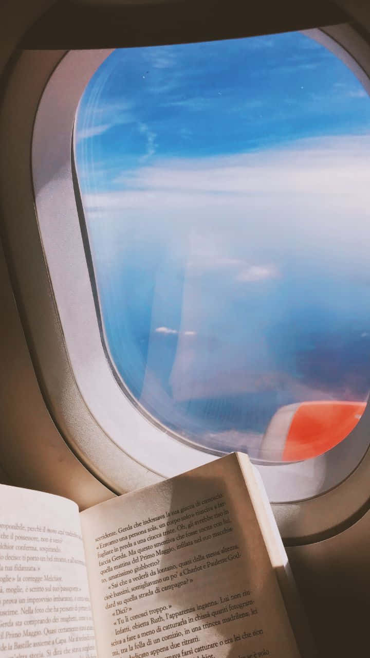 飛機 桌布 壁紙 — 一本書打開放在飛機座位上，窗外雲朵飄過，可見紅白相間的翼尖。