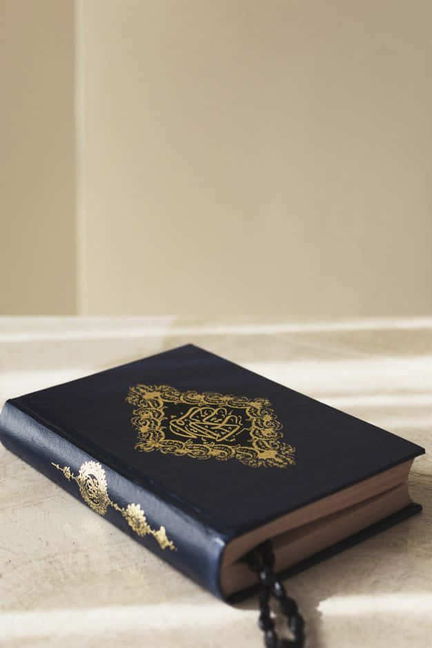 Quran 桌布 壁紙 — 一本深藍色古蘭經，帶有金色浮雕阿拉伯文字和花卉圖案，放置在淺米色表面上，帶有柔和陰影。