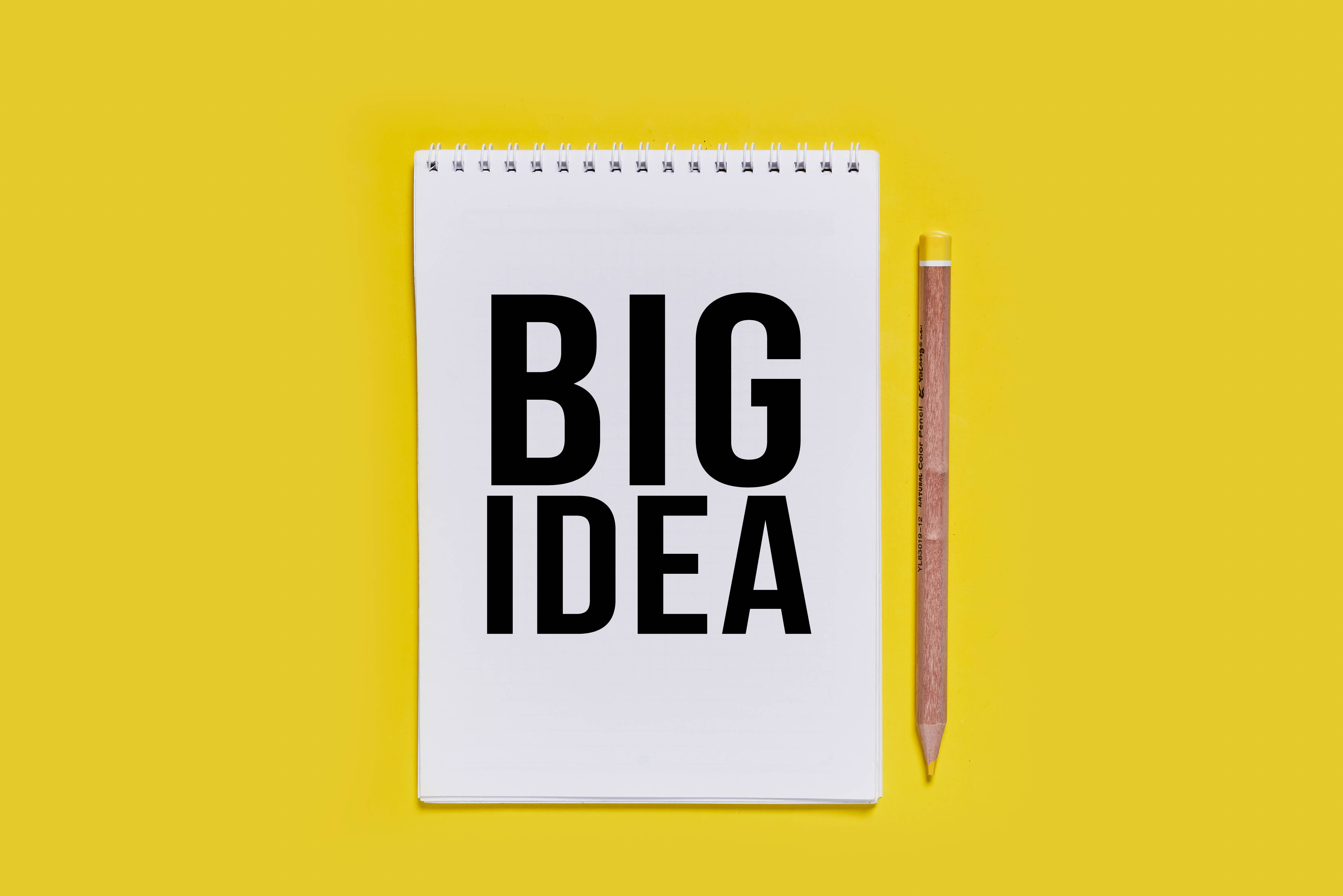 黑色 桌布 壁紙 — 一本白色螺旋筆記本，上面有粗體黑色文字寫著「BIG IDEA」，旁邊放著一支木鉛筆，背景是鮮黃色。