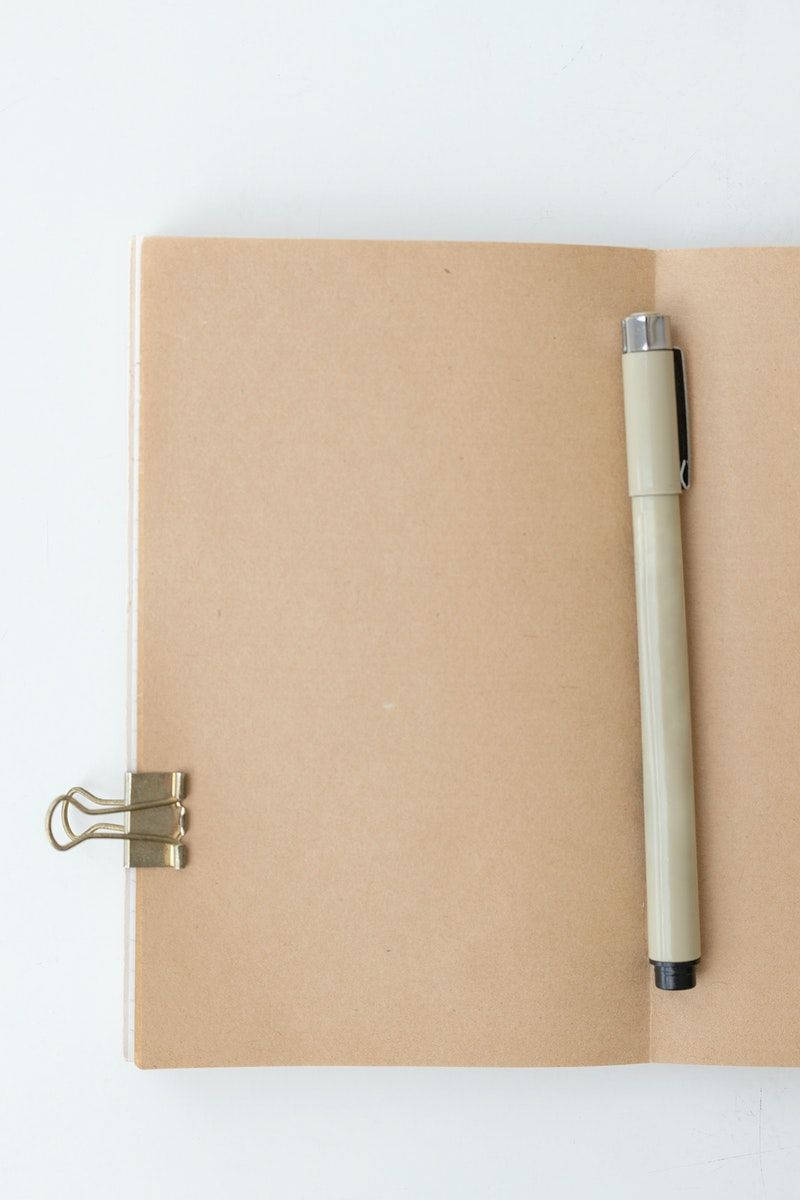 paper 桌布 壁紙 — 一本簡約的米色筆記本，搭配金色活頁夾和奶油色筆，放置在潔白的表面上。