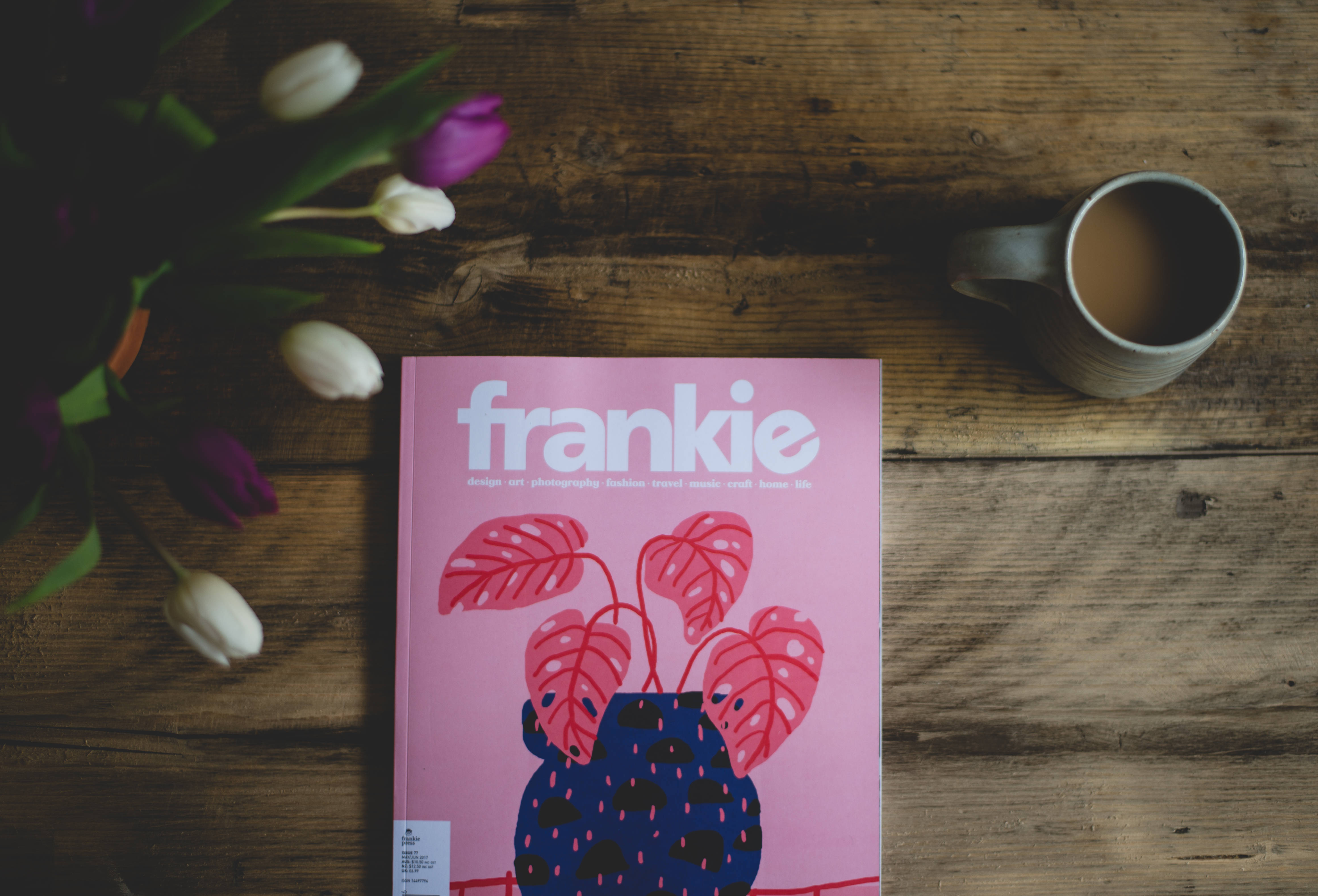 Magazine 桌布 壁紙 — 一本粉紅色雜誌，上面有藍色花瓶和粉紅色葉子，放在木桌上，旁邊有鬱金香和咖啡杯。