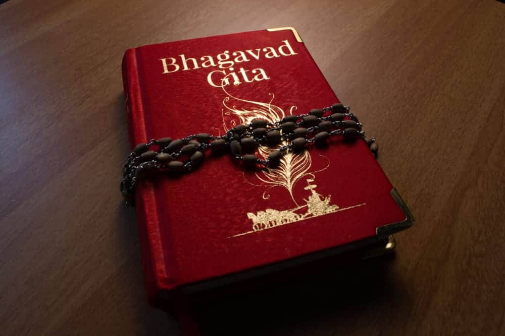 hindu iPhone 桌布 壁紙 — 一本紅色《薄伽梵歌》書籍，帶有金色字體和華麗設計，放在木桌上，並有一串深色珠串的玫瑰經覆蓋在封面上。