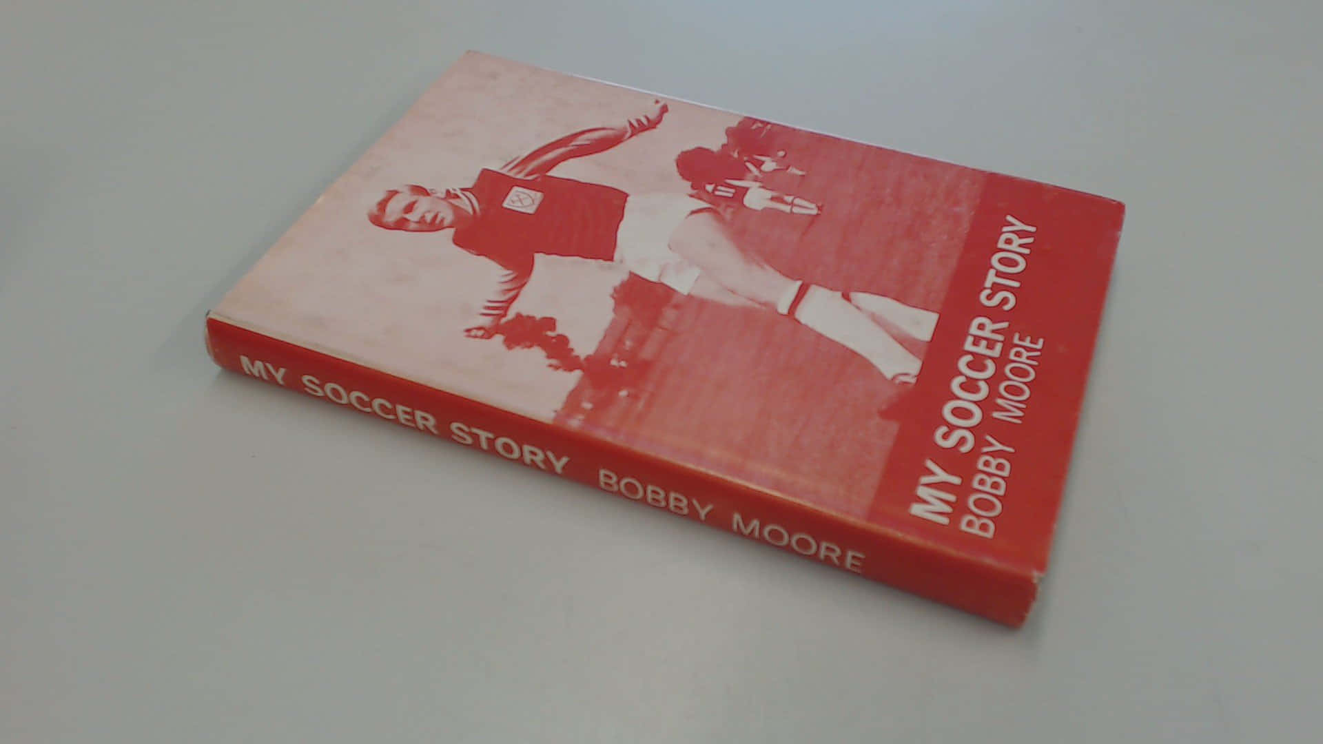書本 桌布 壁紙 — 一本紅色書，名為《My Soccer Story》，作者Bobby Moore，傾斜放在淺灰色平面上。