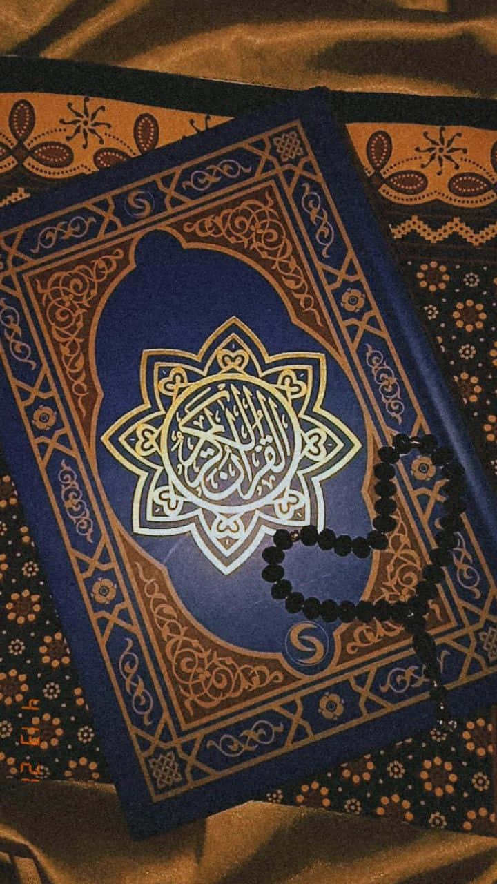 Quran 桌布 壁紙 — 一本華麗的藍色古蘭經，帶有金色書法和精緻邊框，放在圖案布料上，旁邊有一條黑色祈禱珠項鍊。