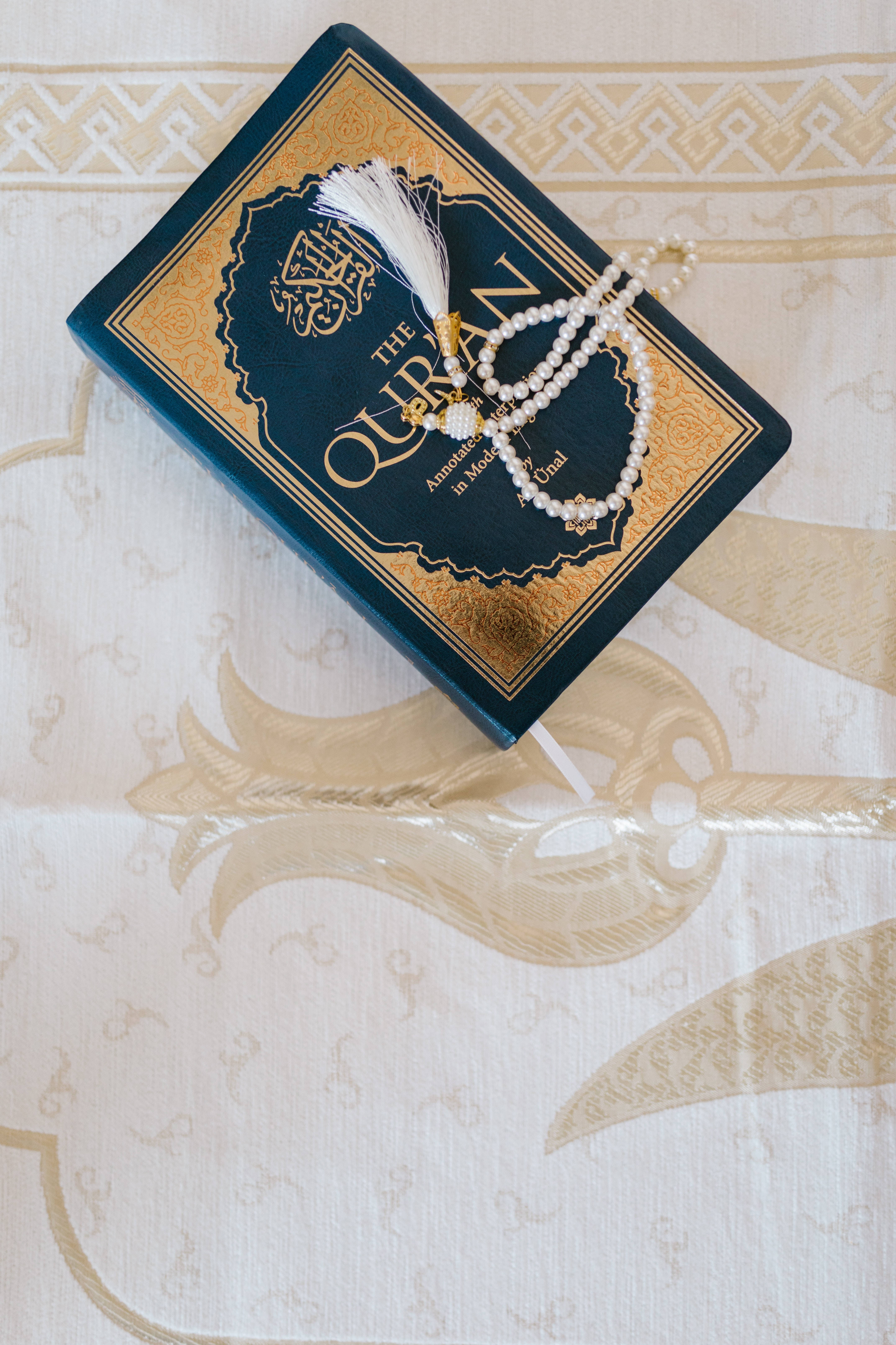 Best Islamic 桌布 壁紙 — 一本藍色和金色的古蘭經，帶有白色流蘇和珍珠念珠，放在帶有金色圖案的華麗奶油色布料上。