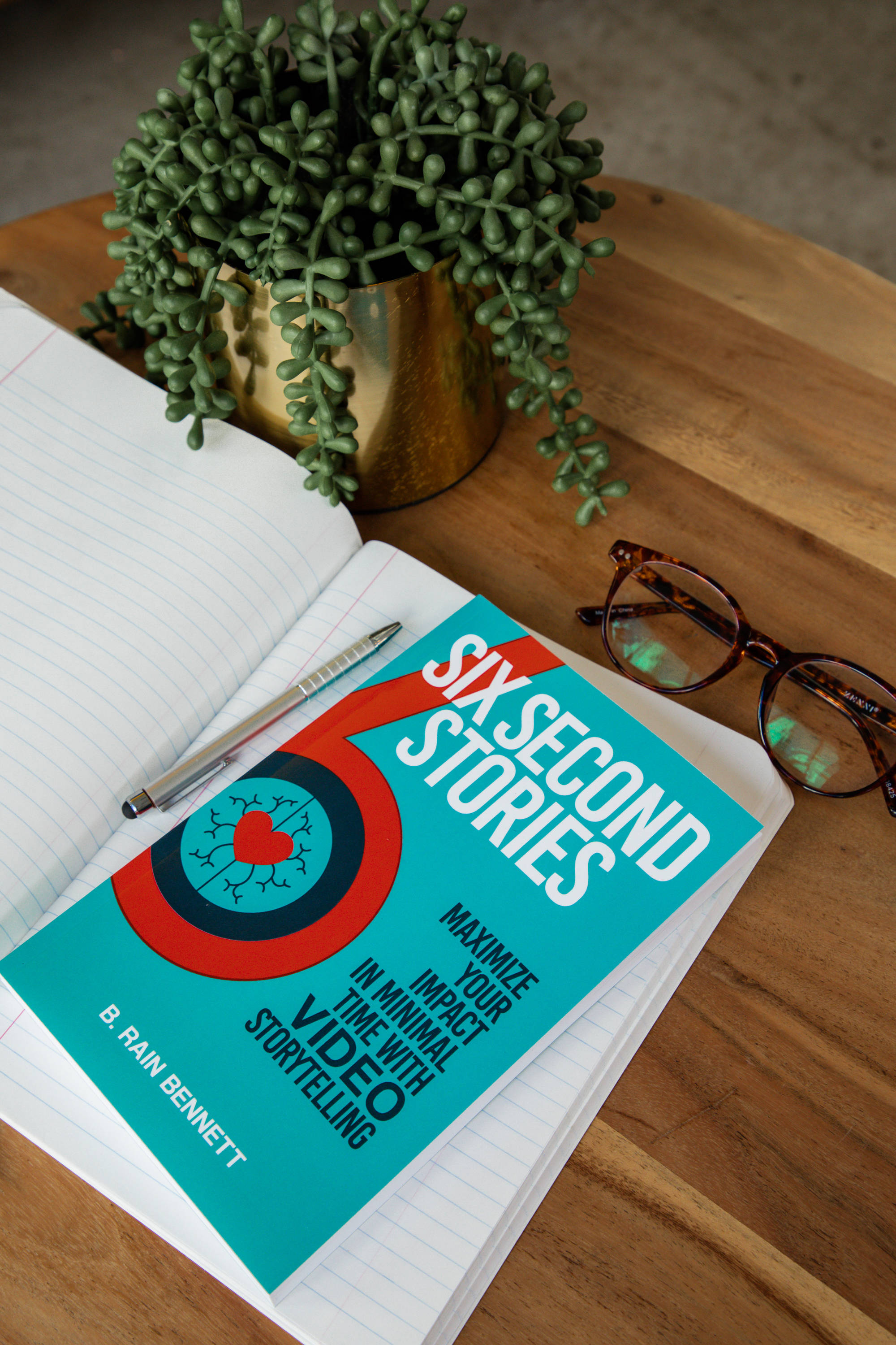 Journals 桌布 壁紙 — 一本青綠色書名為《Six Second Stories》的書放在打開的筆記本上，旁邊有一個金色花盆，裡面有垂下的綠色多肉植物和玳瑁眼鏡，放在木桌上。