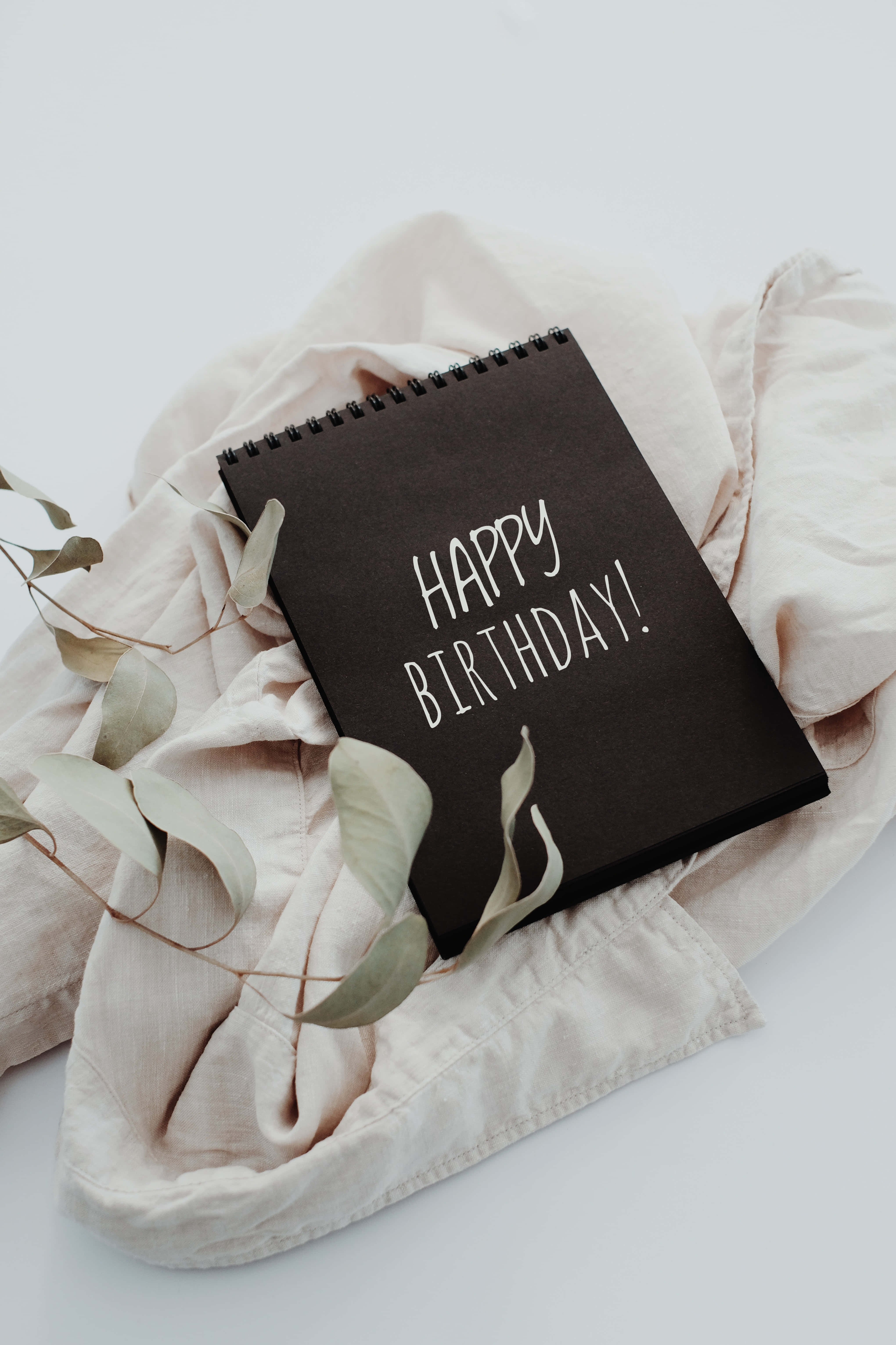 Cute Birthday 桌布 壁紙 — 一本黑色螺旋筆記本，上面有白色手寫字體的「HAPPY BIRTHDAY!」，放在柔軟的米色布料上，搭配乾燥的尤加利葉。
