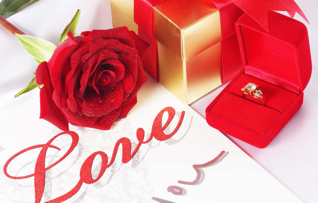 Marriage 桌布 壁紙 — 一朵帶有露珠的鮮紅玫瑰放在一個金色禮物盒和一個打開的紅色天鵝絨戒指盒旁邊，盒內有一枚鑽石戒指，白色表面上帶有閃爍的「Love」字樣。