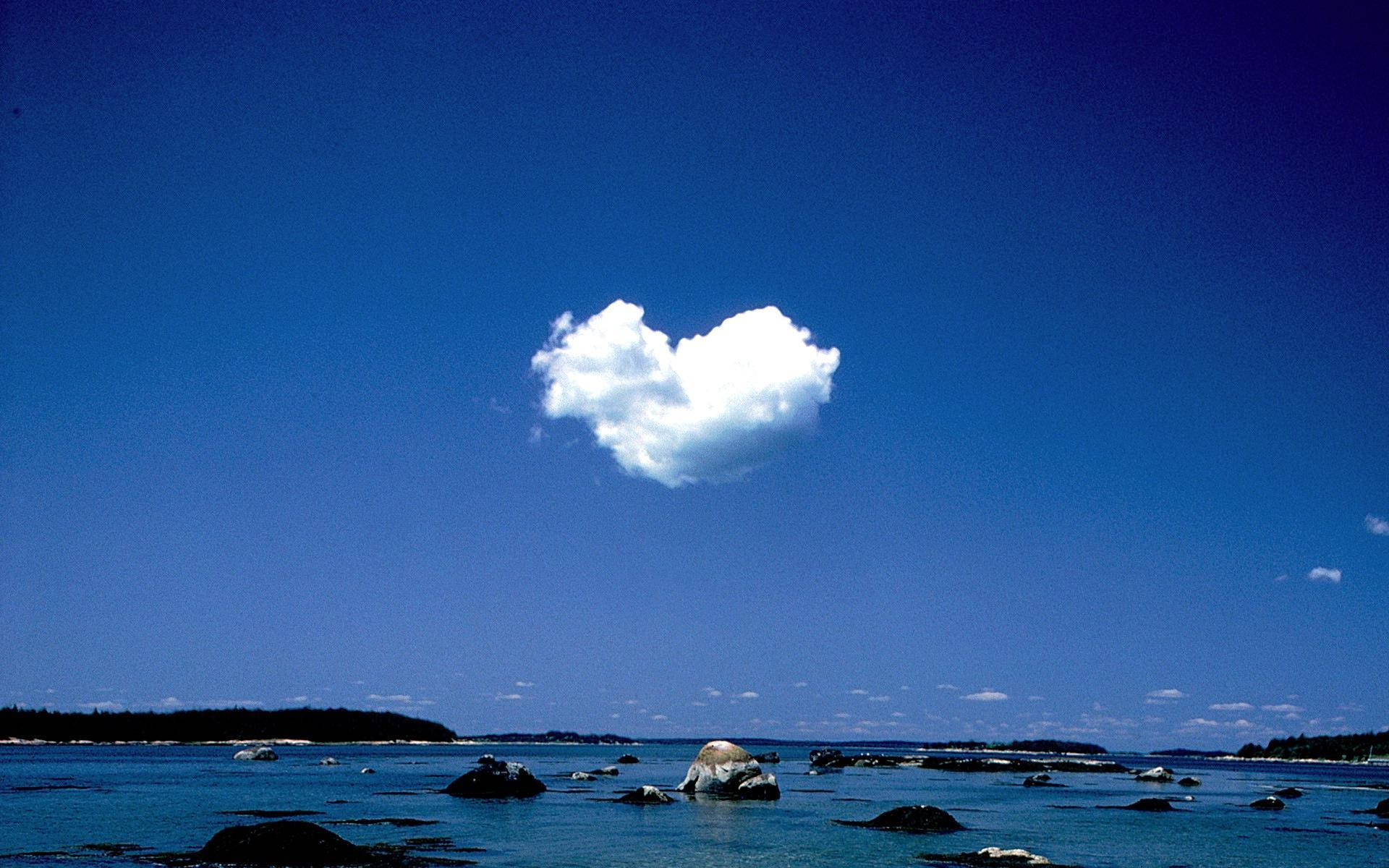 Nature Love 桌布 壁紙 — 一朵心形雲朵平靜地漂浮在平靜的藍色海洋上，海洋點綴著深色岩石和遠處樹木成行的海岸。