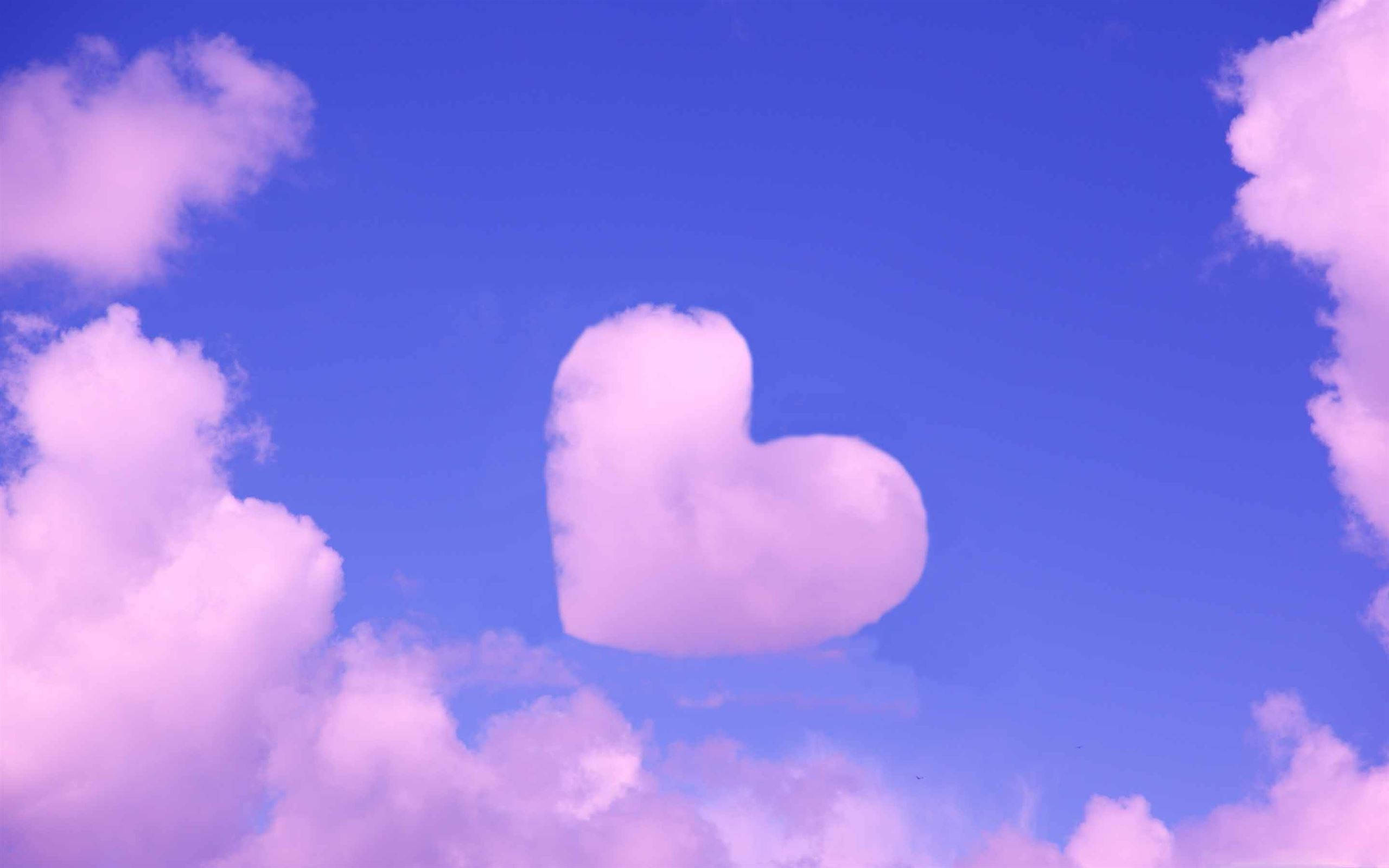 Pastel Pink Heart 桌布 壁紙 — 一朵柔軟的粉紅色心形雲朵在鮮豔的藍天中寧靜地漂浮，周圍環繞著蓬鬆的粉彩雲朵。