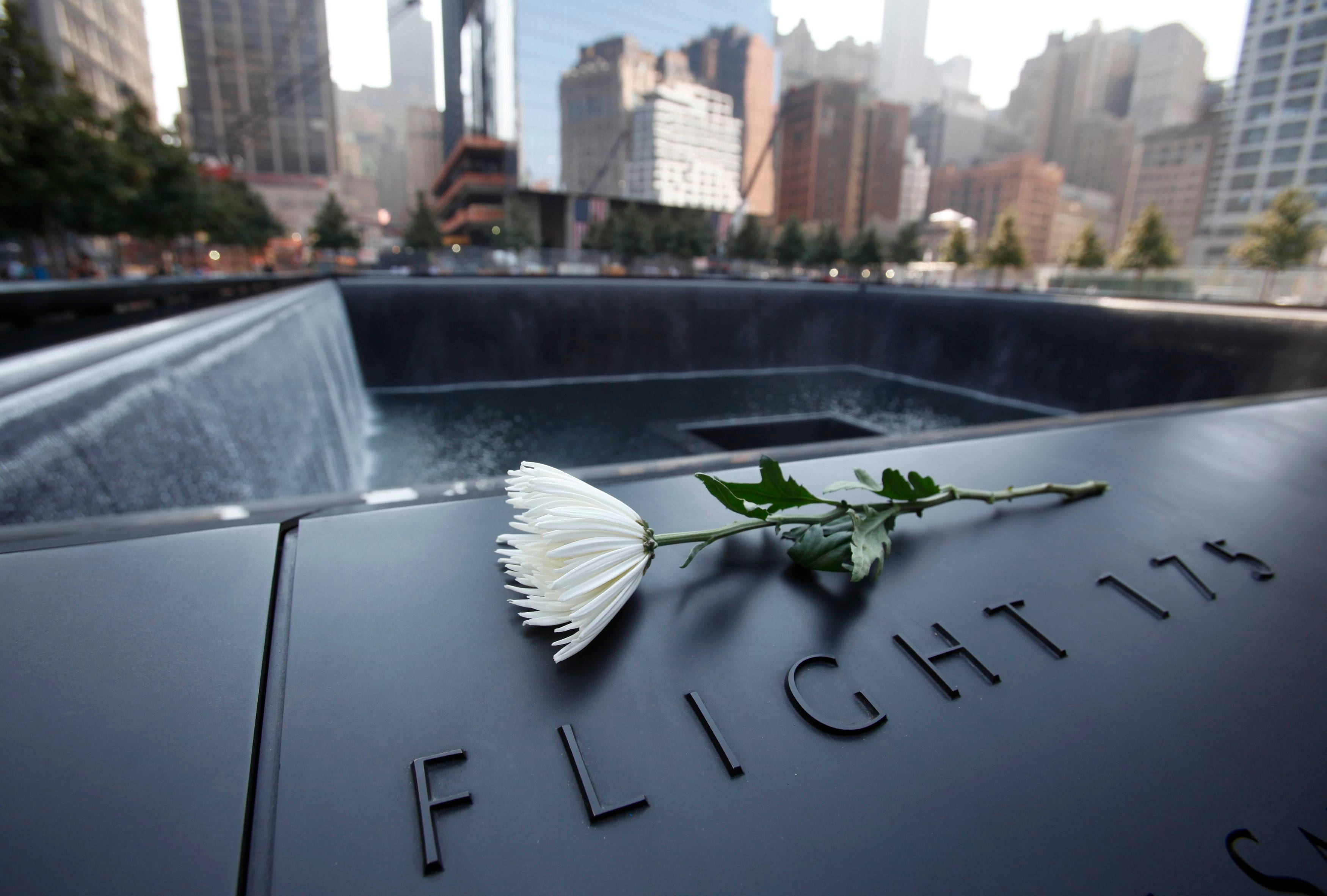 911 Memorial 桌布 壁紙 — 一朵白色鮮花莊嚴地放在刻有 'FLIGHT 175' 的黑色紀念牌上，旁邊是城市景觀中的層疊水景。