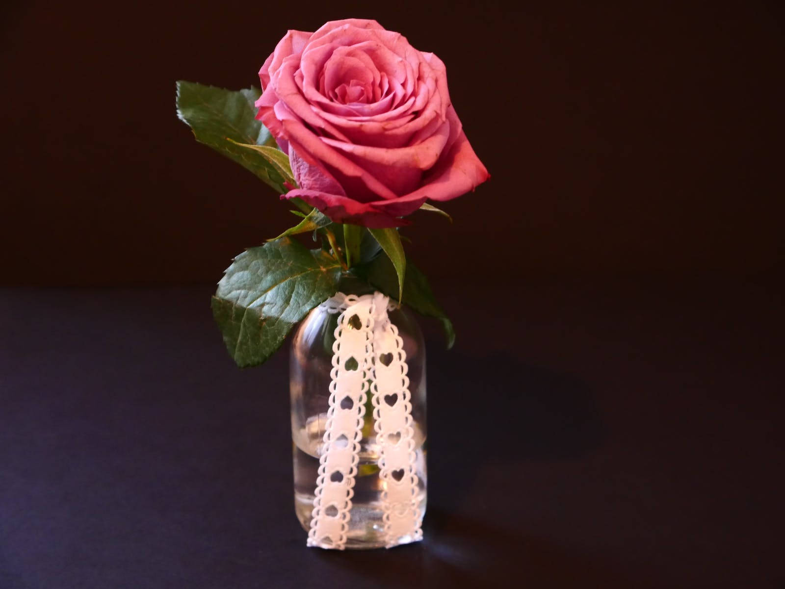 Rose HD 桌布 壁紙 — 一朵粉紅玫瑰放在透明玻璃花瓶，飾以白色心形蕾絲絲帶，背景深色。