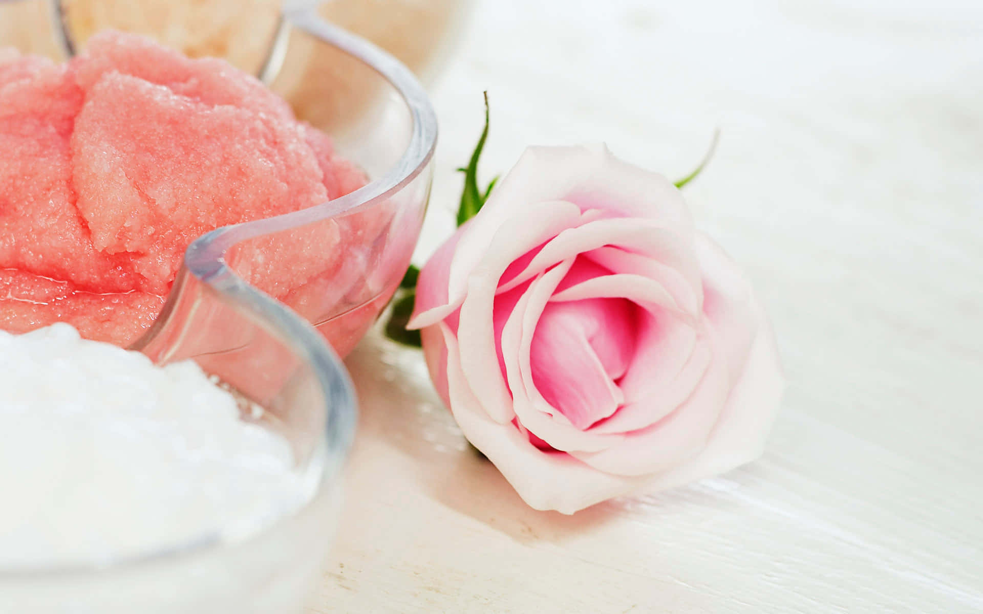 Spa Treatments 桌布 壁紙 — 一朵精緻的粉紅玫瑰放在明亮的白色木質表面上，旁邊是裝有粉紅和白色糖磨砂的玻璃碗。