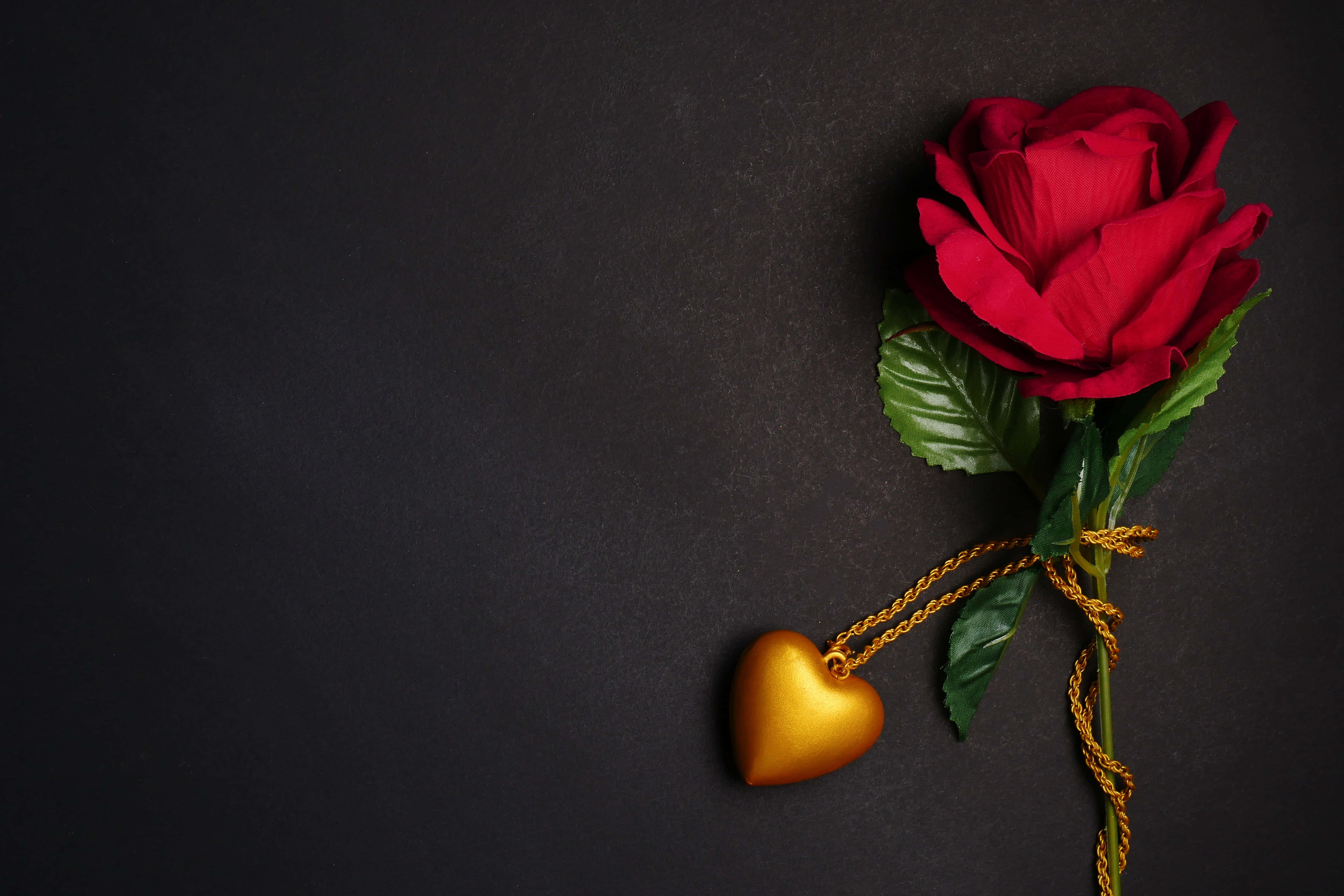 Red Rose 桌布 壁紙 — 一朵紅玫瑰，帶有綠葉和一個用金繩繫著的金色心形吊墜，放置在深色紋理表面上。