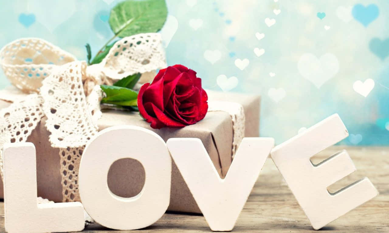 1280x768 Rose 桌布 壁紙 — 一朵紅玫瑰和白色「LOVE」字母放在一個用棕色紙和蕾絲包裹的禮物上，背景是心形圖案的藍色。