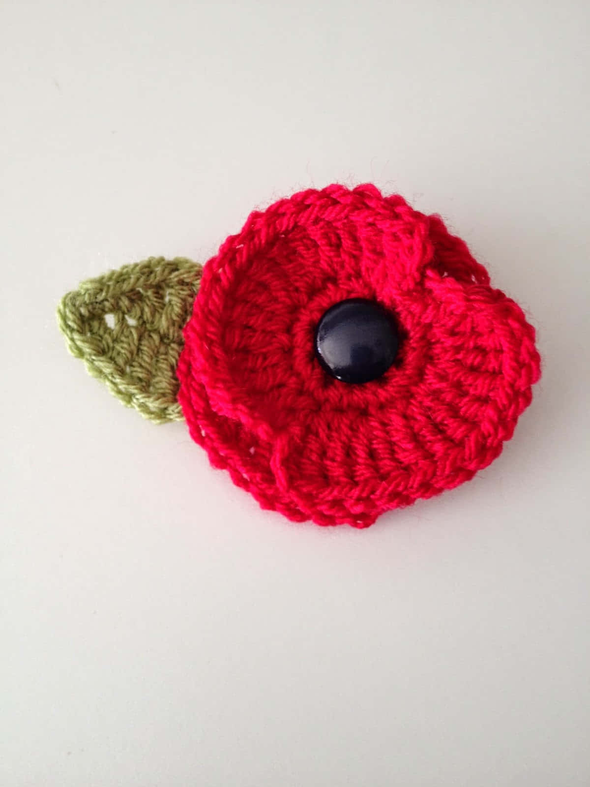 Crochet 桌布 壁紙 — 一朵鮮紅色鉤針編織的罌粟花，帶有黑色鈕扣中心和綠色葉子，放在白色平面上。