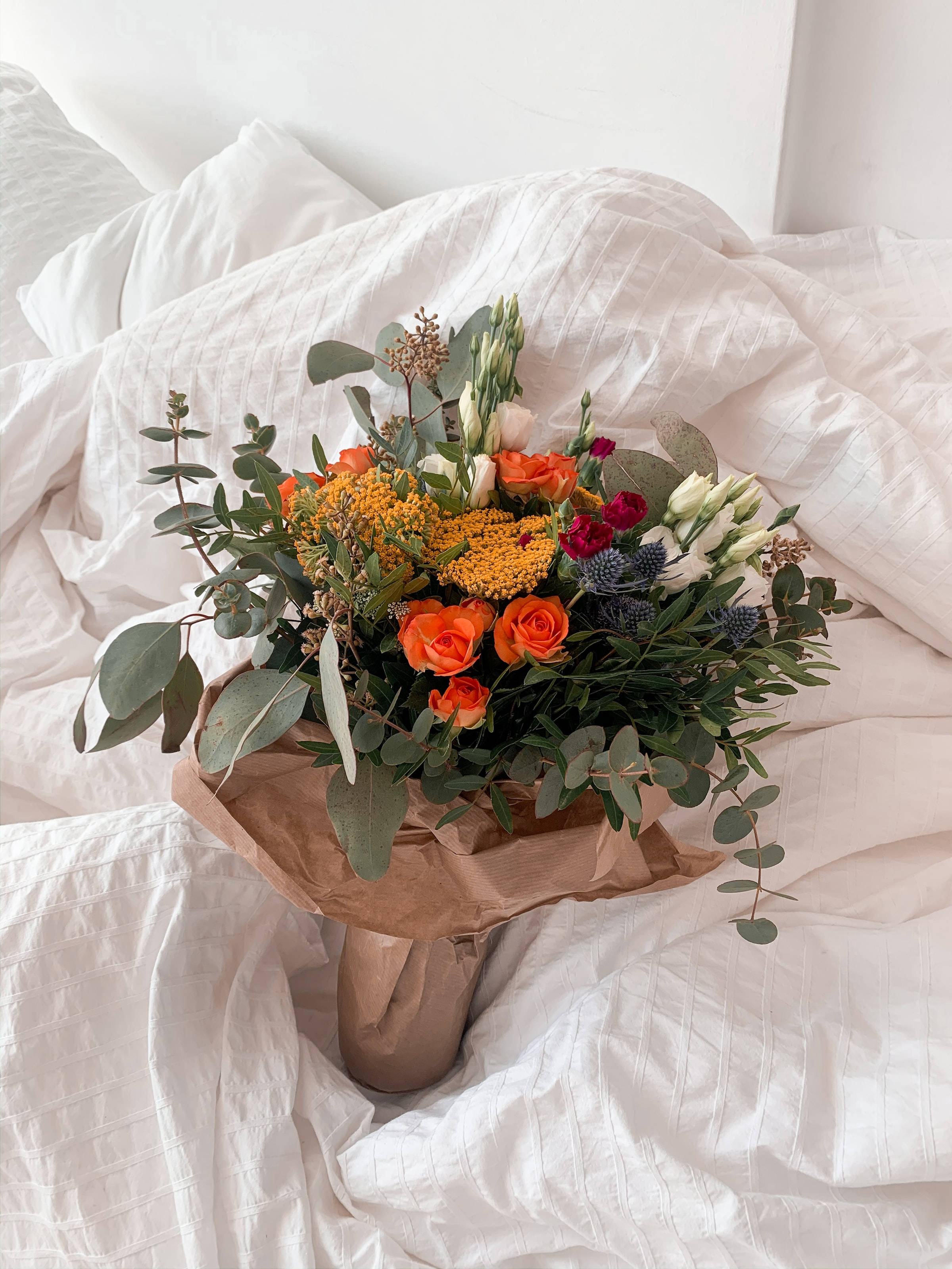 Bouquet 桌布 壁紙 — 一束充滿活力的橙色玫瑰、黃色花朵和綠色植物，用棕色紙包裹，放在皺巴巴的白色床罩上。