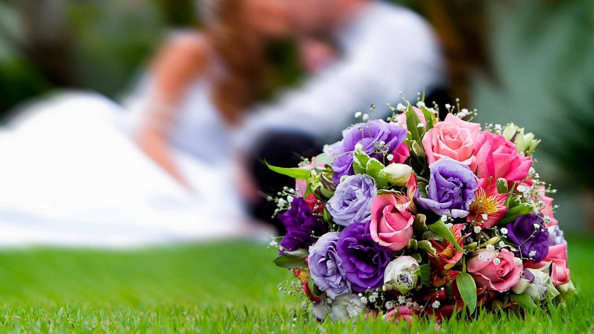 Wedding 桌布 壁紙 — 一束充滿活力的粉紅色和紫色玫瑰放在茂密的綠草地上，背景有一對模糊的情侶在接吻。