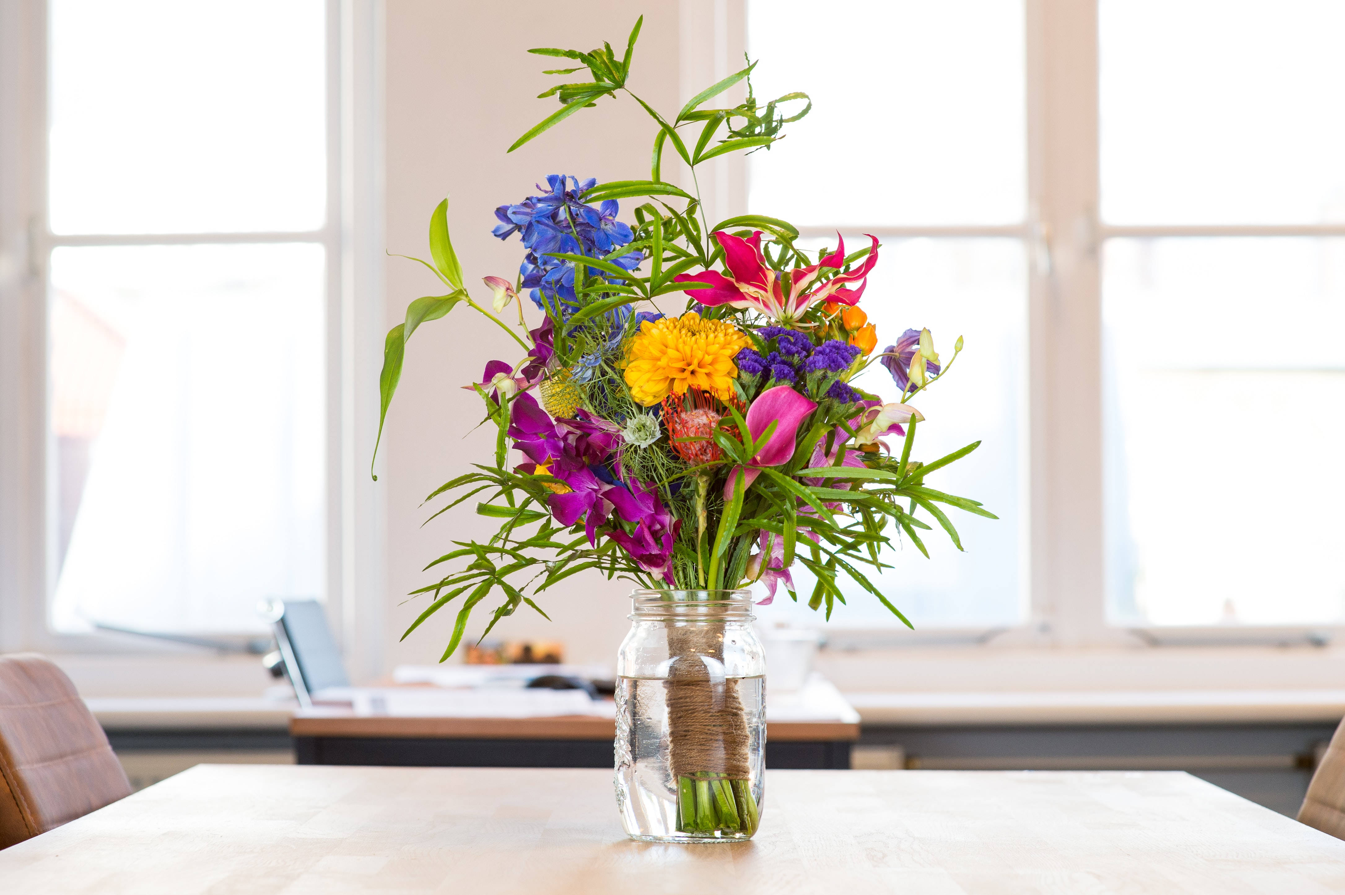 Flowers 桌布 壁紙 — 一束充滿活力的藍色、粉紅色、黃色和紫色花朵放在玻璃罐中，置於明亮窗戶附近的木桌上。