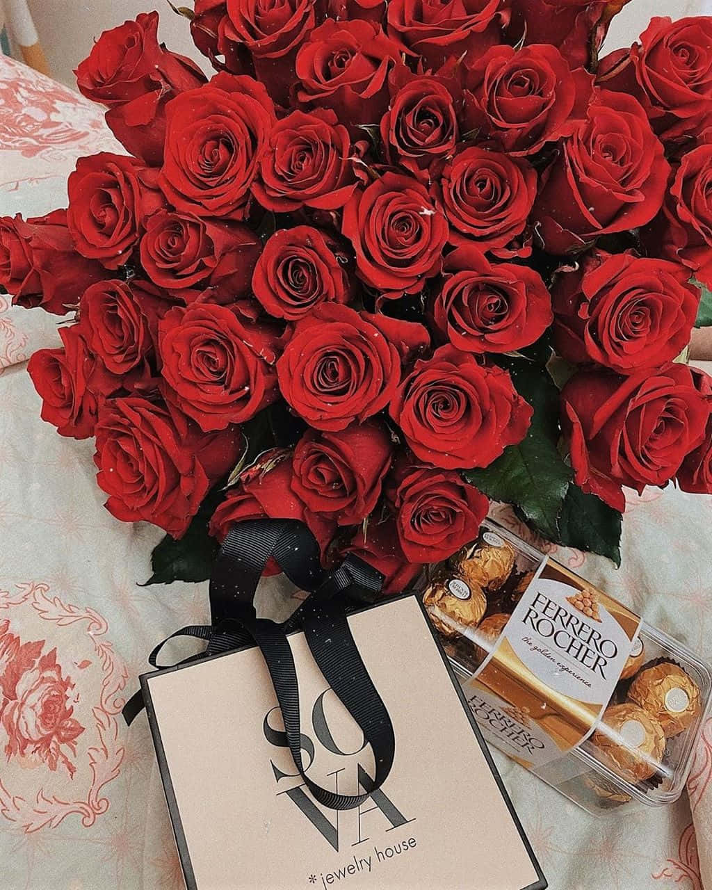 情人節 桌布 壁紙 — 一束奢華的深紅玫瑰放在花床罩上，旁邊有一個Ferrero Rocher盒子和一個珠寶品牌的禮品袋。