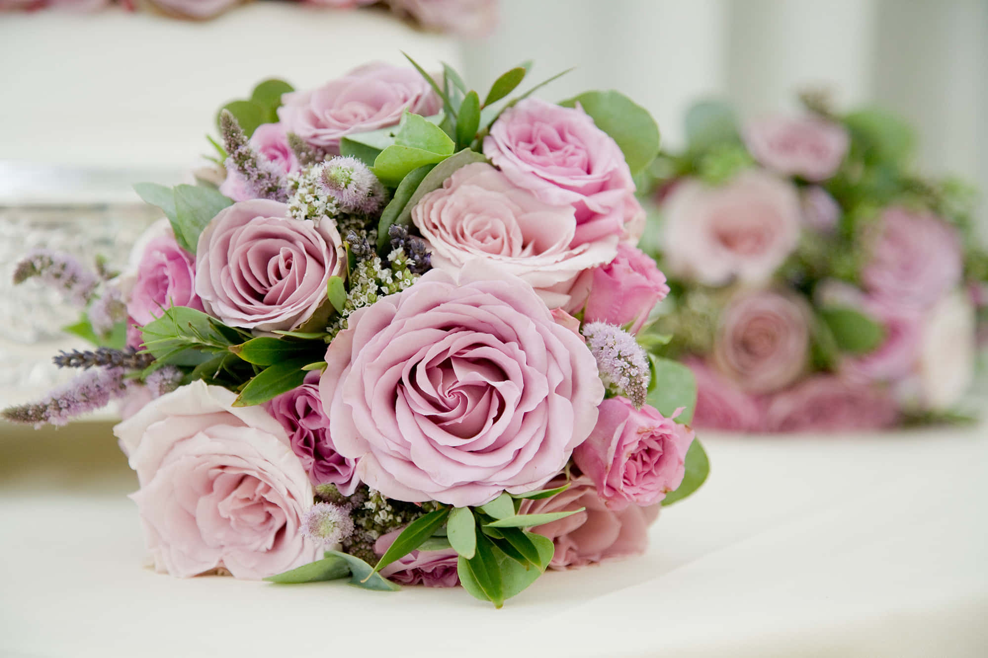 Wedding Bouquet 桌布 壁紙 — 一束柔和焦點的灰粉色玫瑰和綠葉優雅地放在白色表面上，背景中有一個模糊的第二束花。