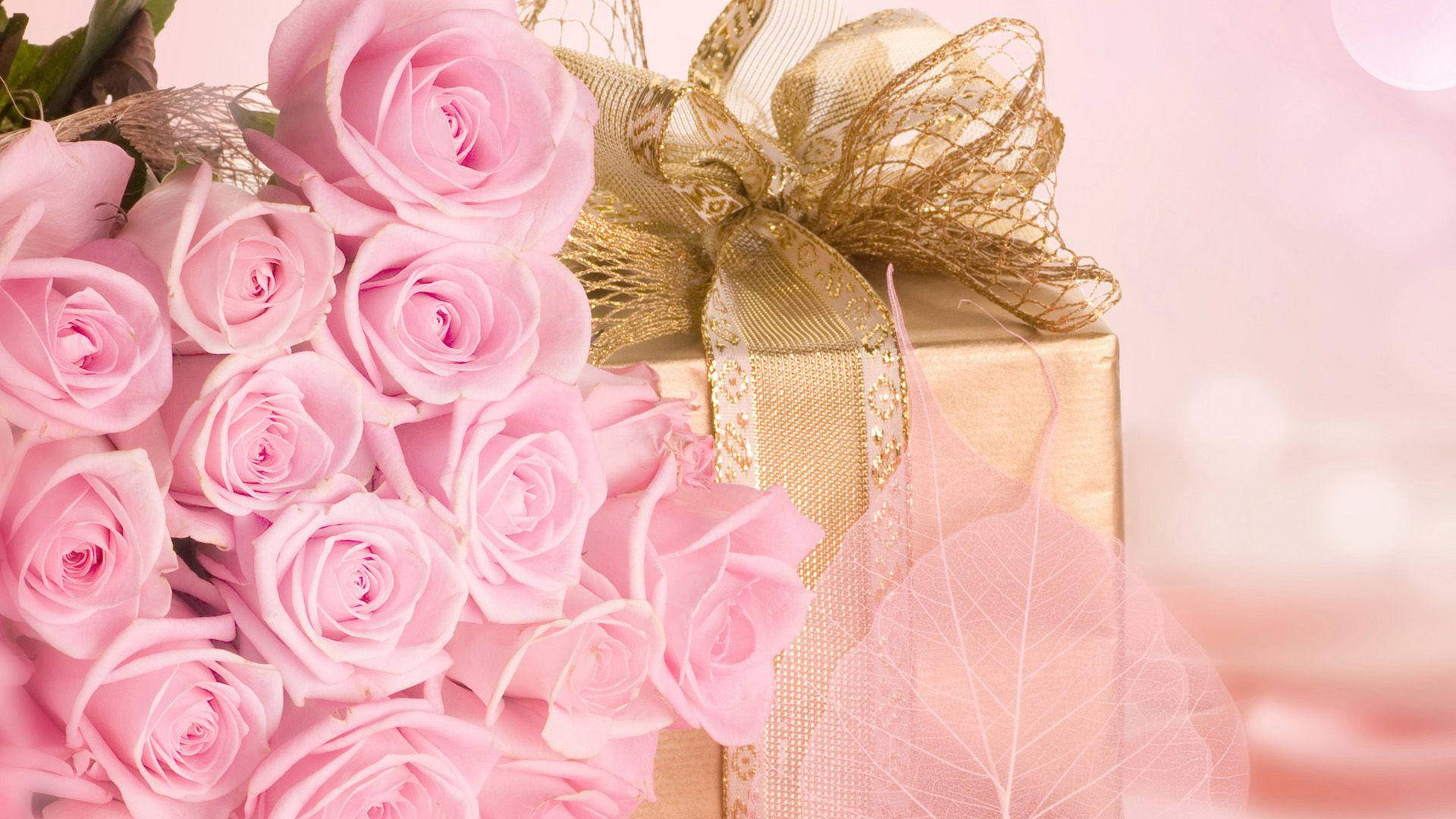 Happy Birthday Flower 桌布 壁紙 — 一束柔和的粉紅玫瑰旁邊有一個金色禮物盒，繫著閃爍的金色蝴蝶結，背景是夢幻般的粉紅色。