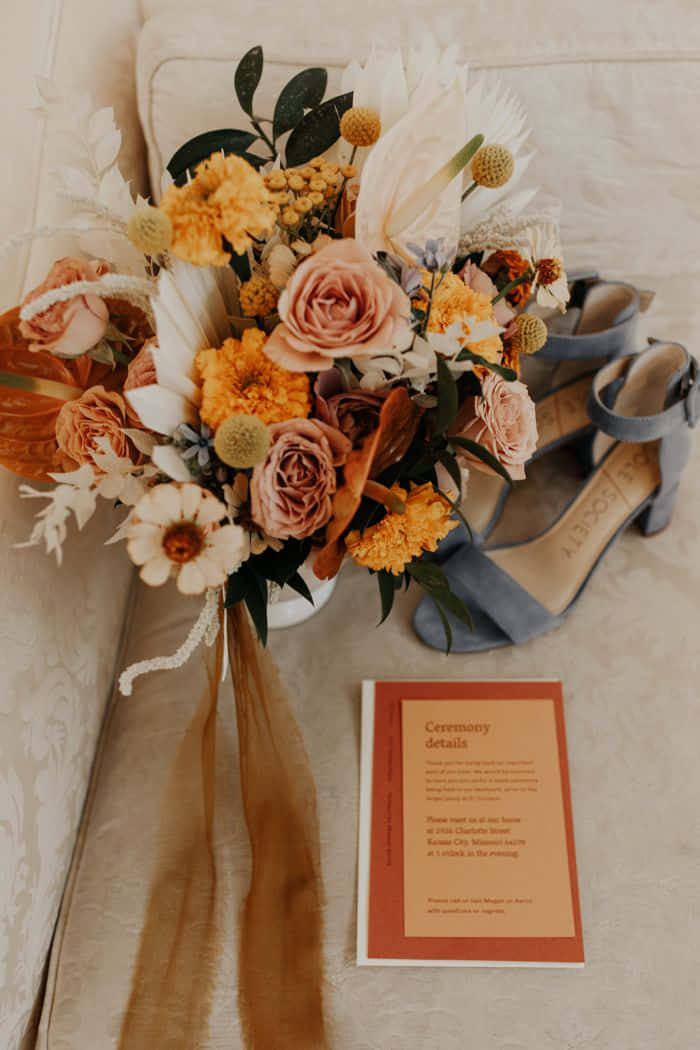 Roses Wedding 桌布 壁紙 — 一束柔和粉彩的玫瑰和乾花，旁邊放著灰藍色高跟鞋和一張婚禮儀式卡，放在有紋理的奶油色沙發上。