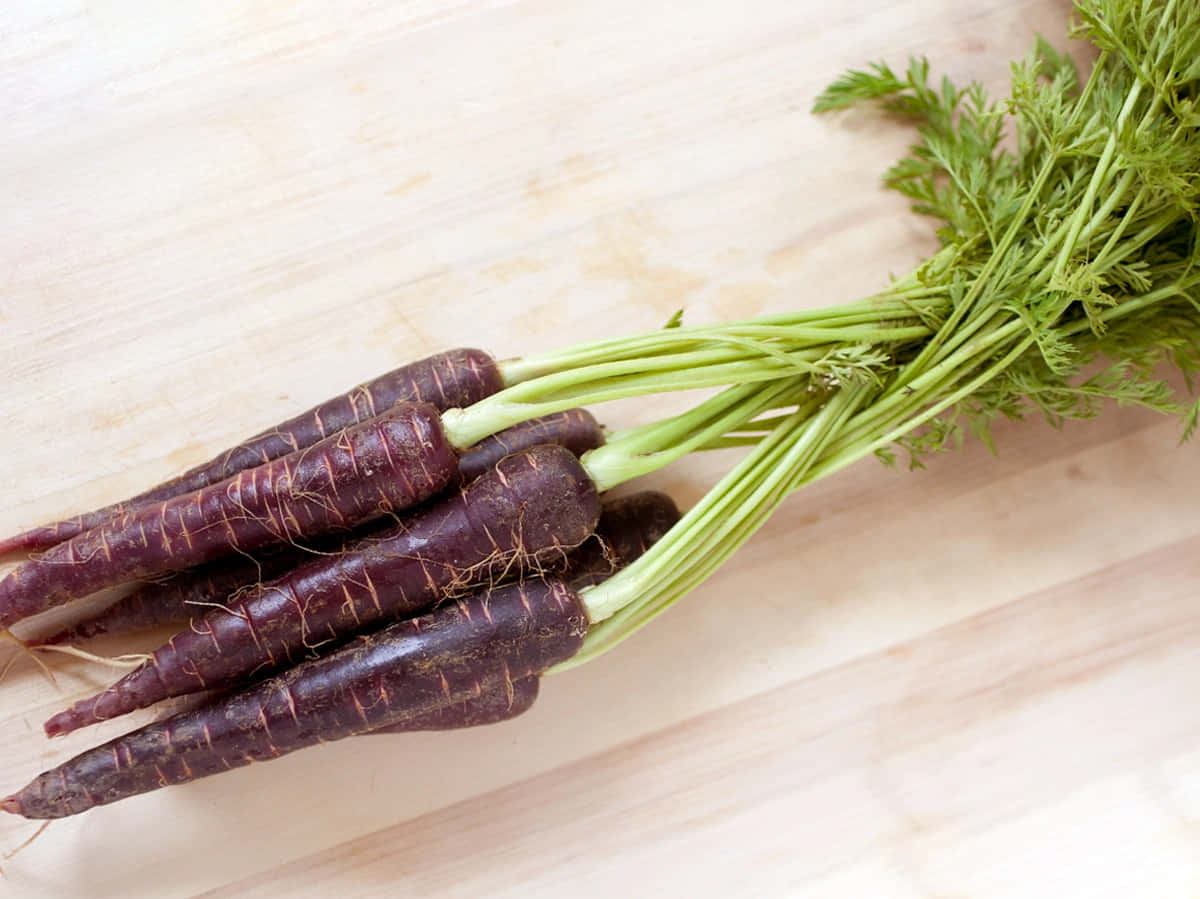Purple Carrots 桌布 壁紙 — 一束深紫色的胡蘿蔔，帶有新鮮的綠色頂部，放在淺色木砧板上。