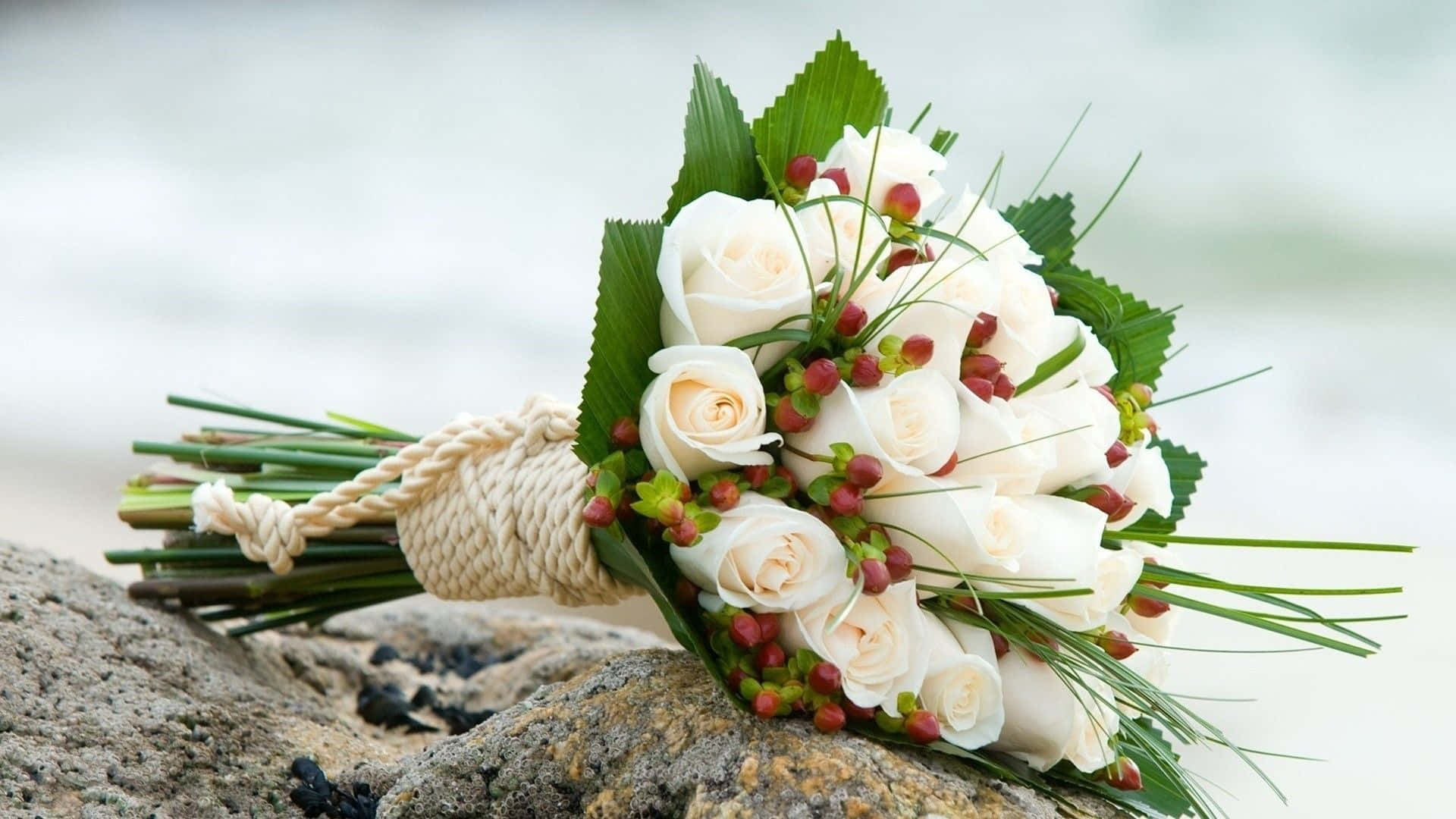 Wedding Flowers 桌布 壁紙 — 一束由白玫瑰和紅莓組成的新娘花束，擱在海邊一塊風化的岩石上。
