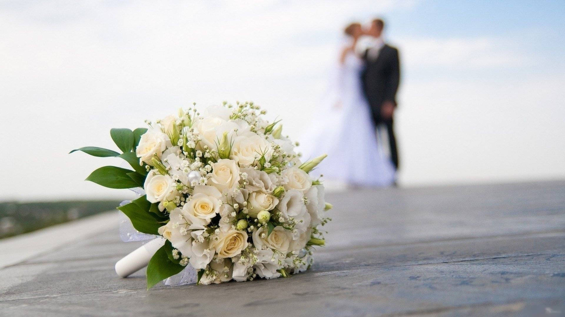 Wedding 桌布 壁紙 — 一束精緻的白玫瑰放在木質表面上，背景中有一對模糊的新郎新娘在接吻。