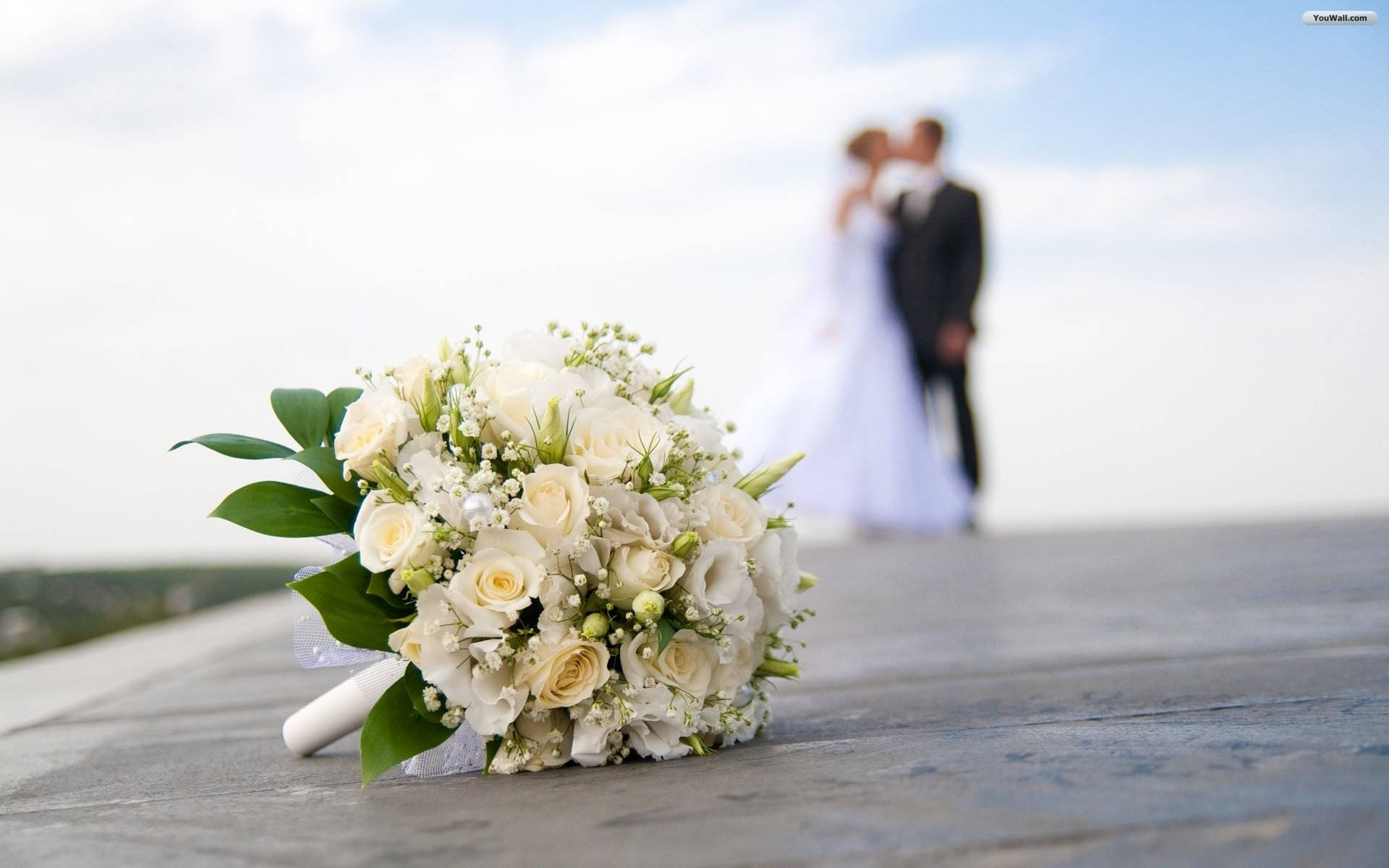 Bridal 桌布 壁紙 — 一束精緻的白色新娘花束放在石頭上，背景中一對模糊的情侶在明亮的天空下接吻。