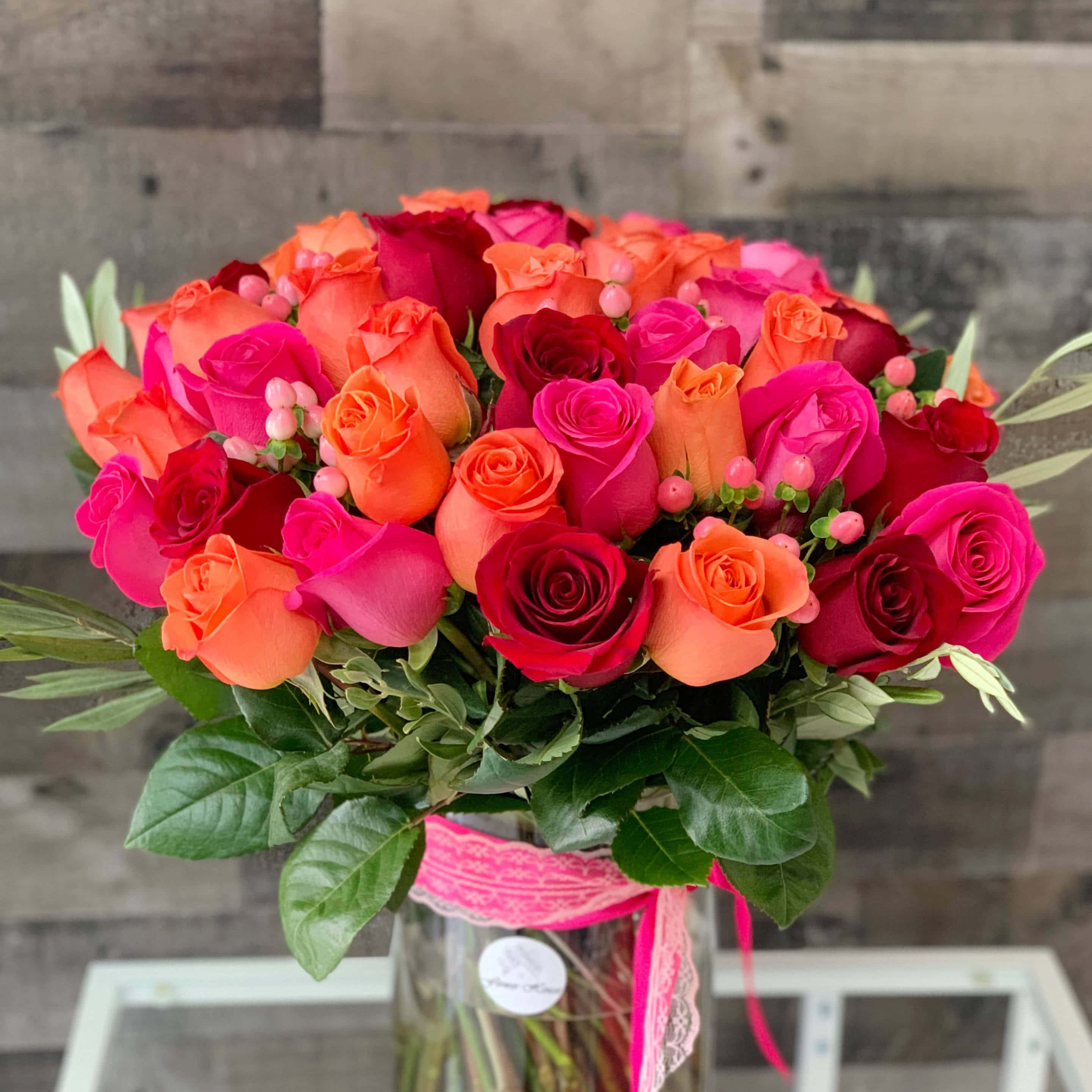 Rose Flower 桌布 壁紙 — 一束鮮豔的紅、橙、粉紅玫瑰，搭配綠葉和粉紅絲帶，置於玻璃花瓶中，背景為木牆。